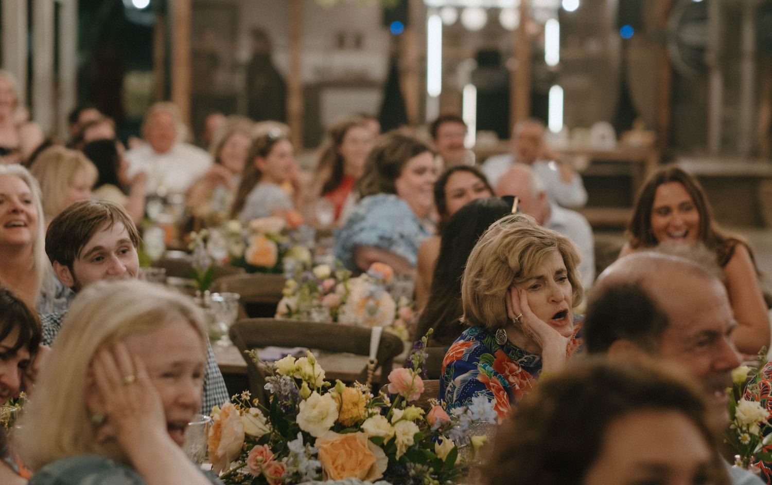Wedding guests laugh and react during speeches at a beautifully decorated indoor reception with floral centerpieces.