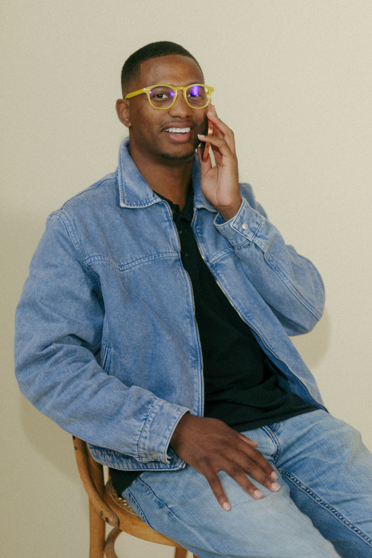 Smiling Black man in denim jacket and yellow glasses talks on smartphone while seated on wooden chair.