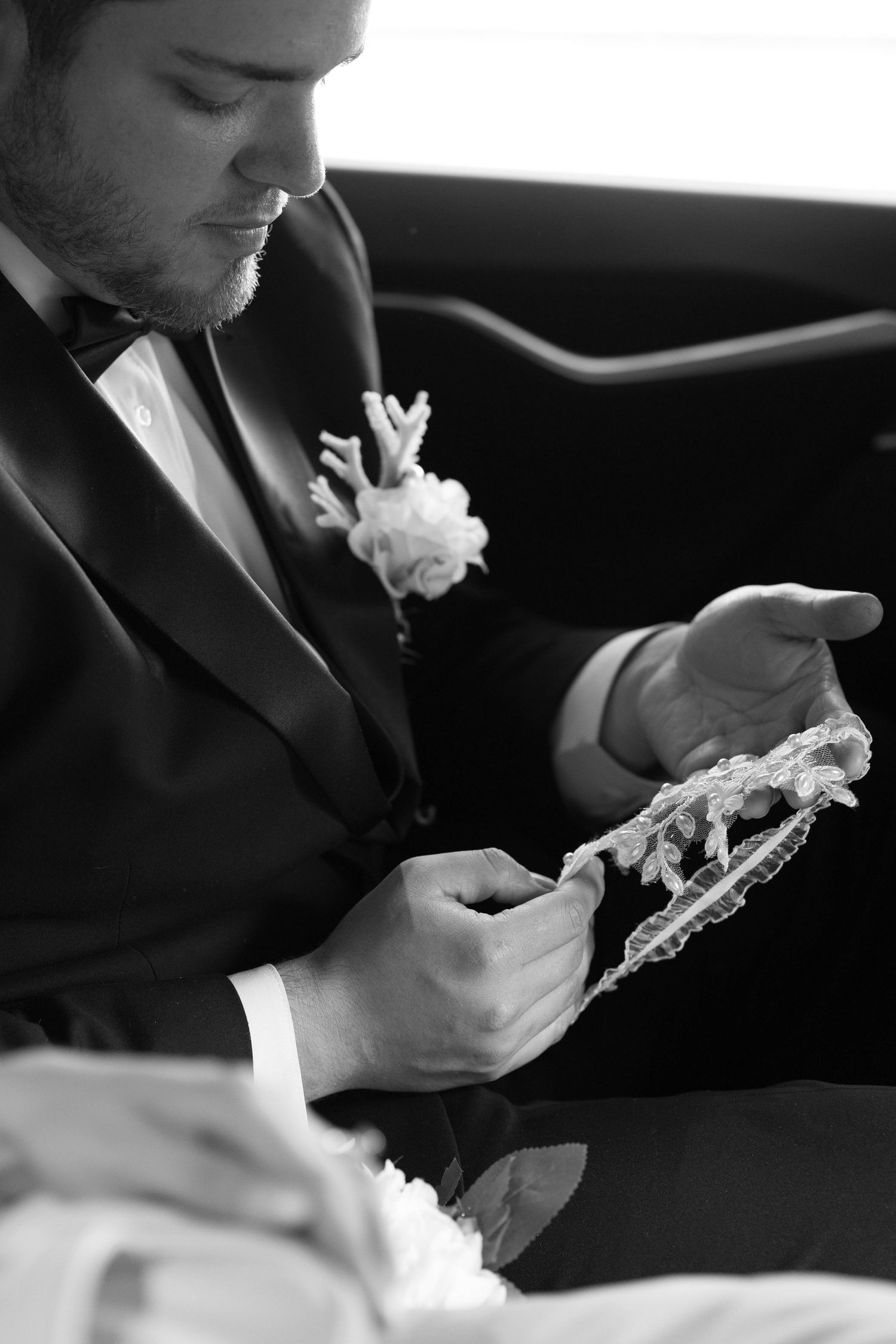 Groom in black tuxedo examines delicate lace bridal accessory while seated in a car.