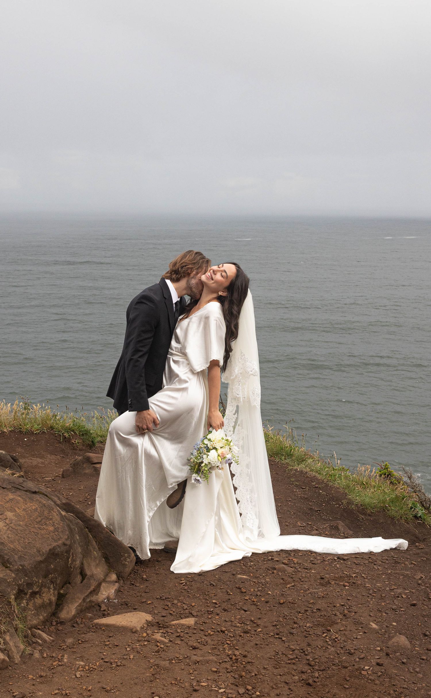 Bride and groom share a romantic kiss on a coastal cliff, bride holding white bouquet in flowing wedding dress with veil.