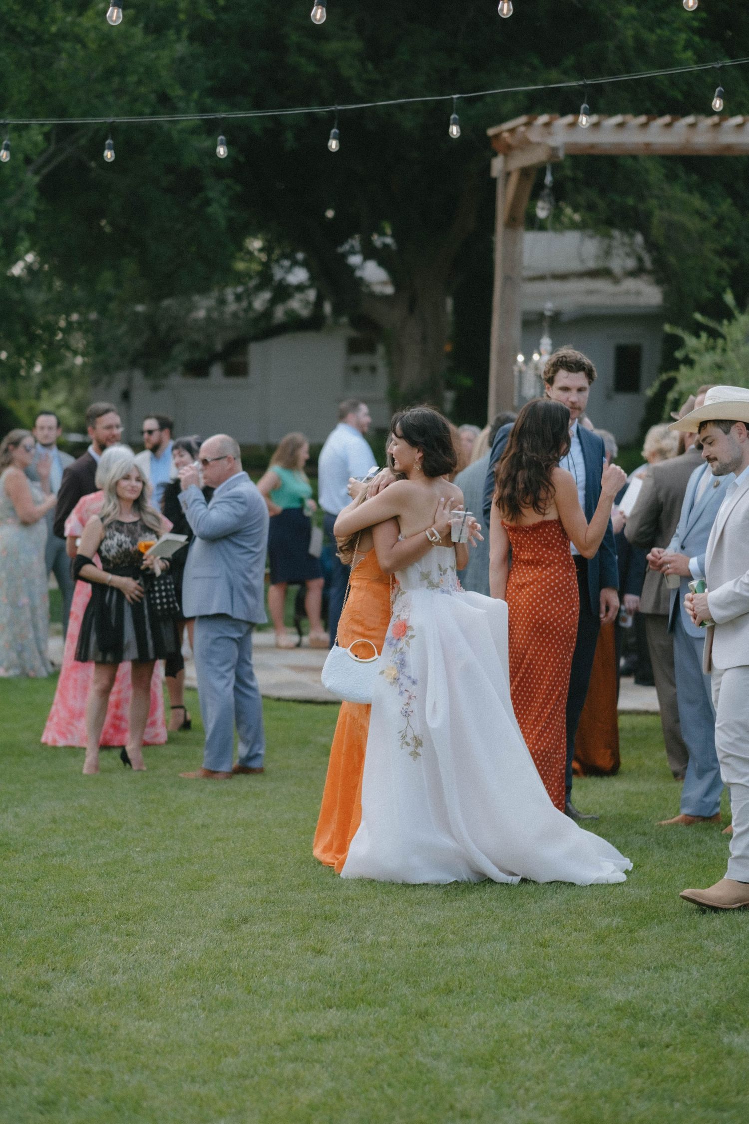 Bride in a floral gown shares a warm embrace with a guest in an orange dress outdoors.