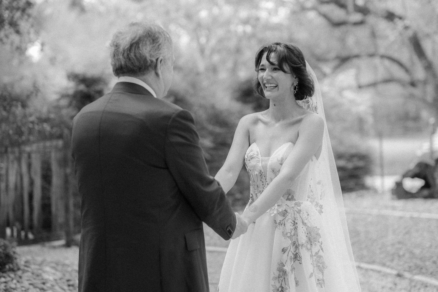 Black and white photo of a smiling bride in a floral gown holding hands with a man outdoors.