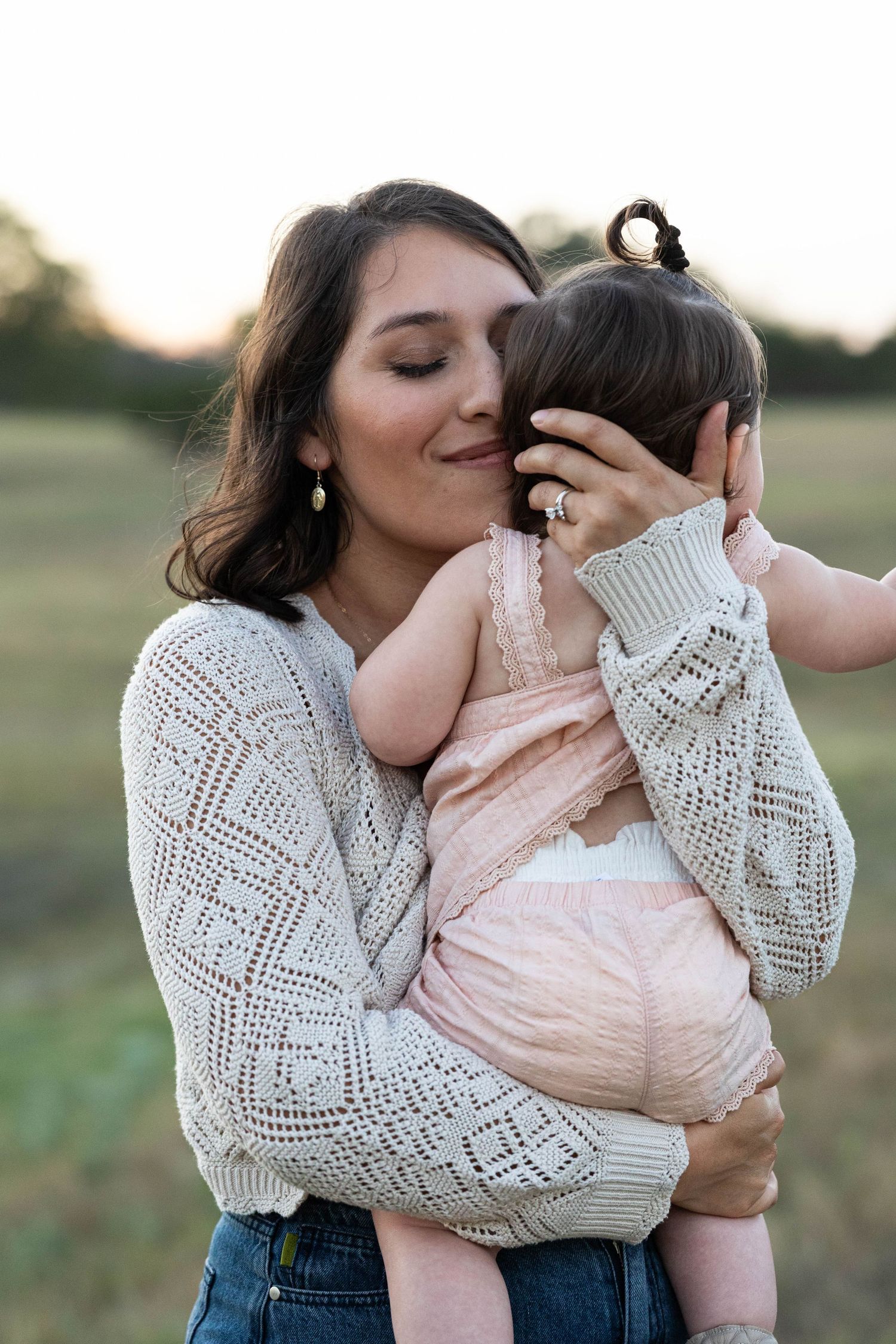 A tender moment as a dark-haired mother in a white knit sweater kisses her baby girl in a field.