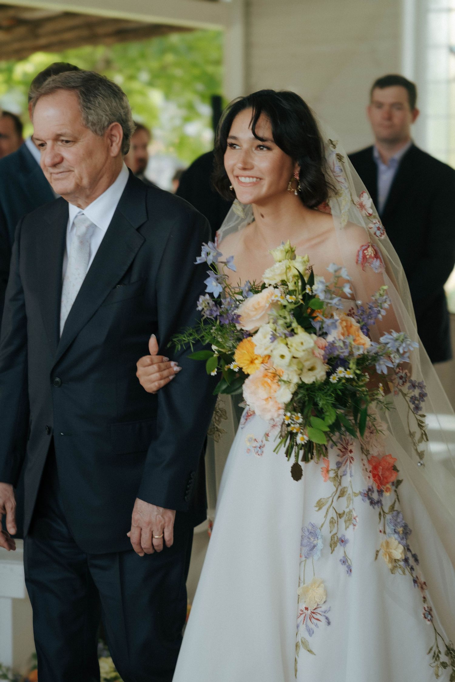 Smiling bride in floral gown walks down aisle with father holding colorful bouquet.