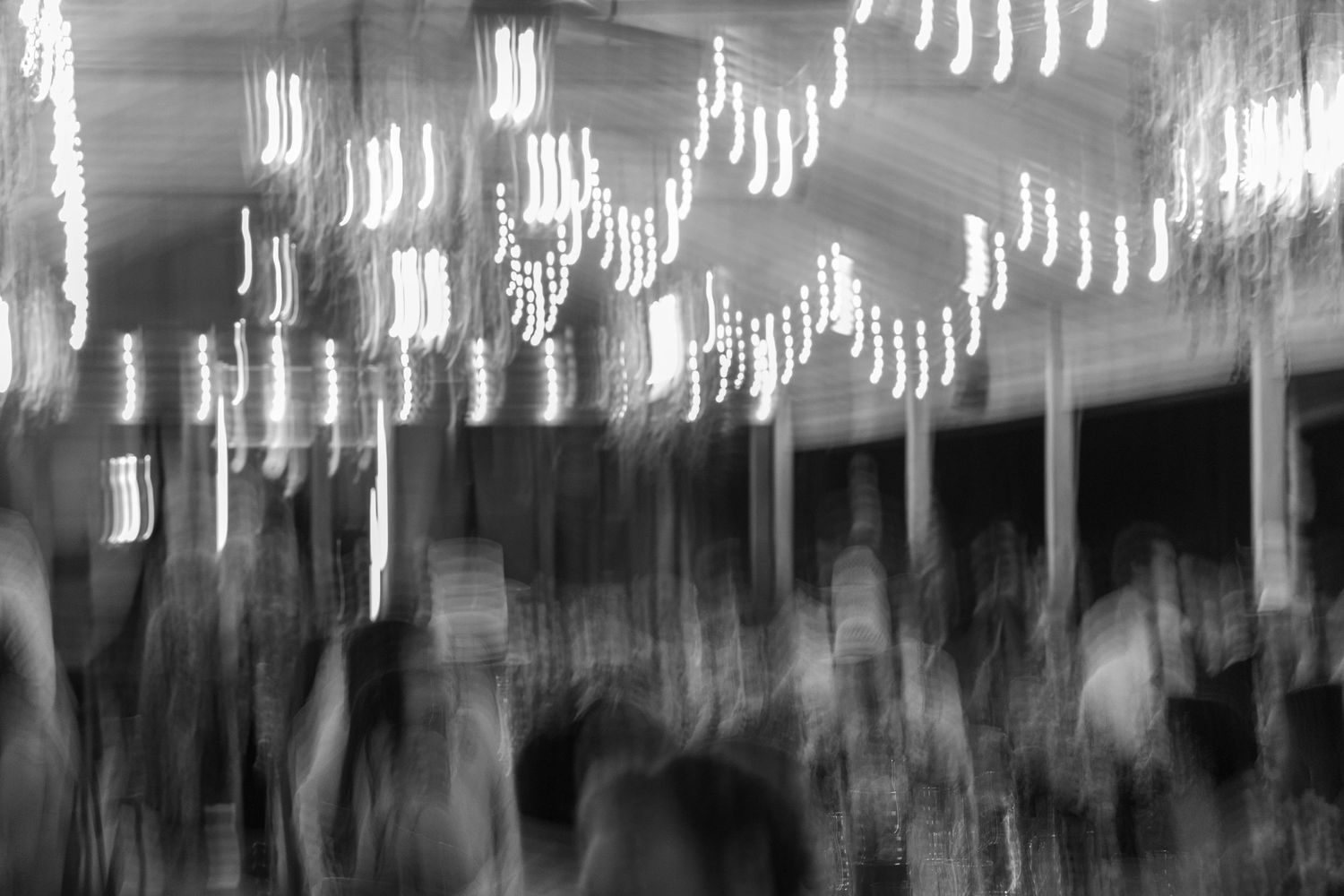 Blurred black and white photo of wedding guests dancing under string lights at a reception venue.