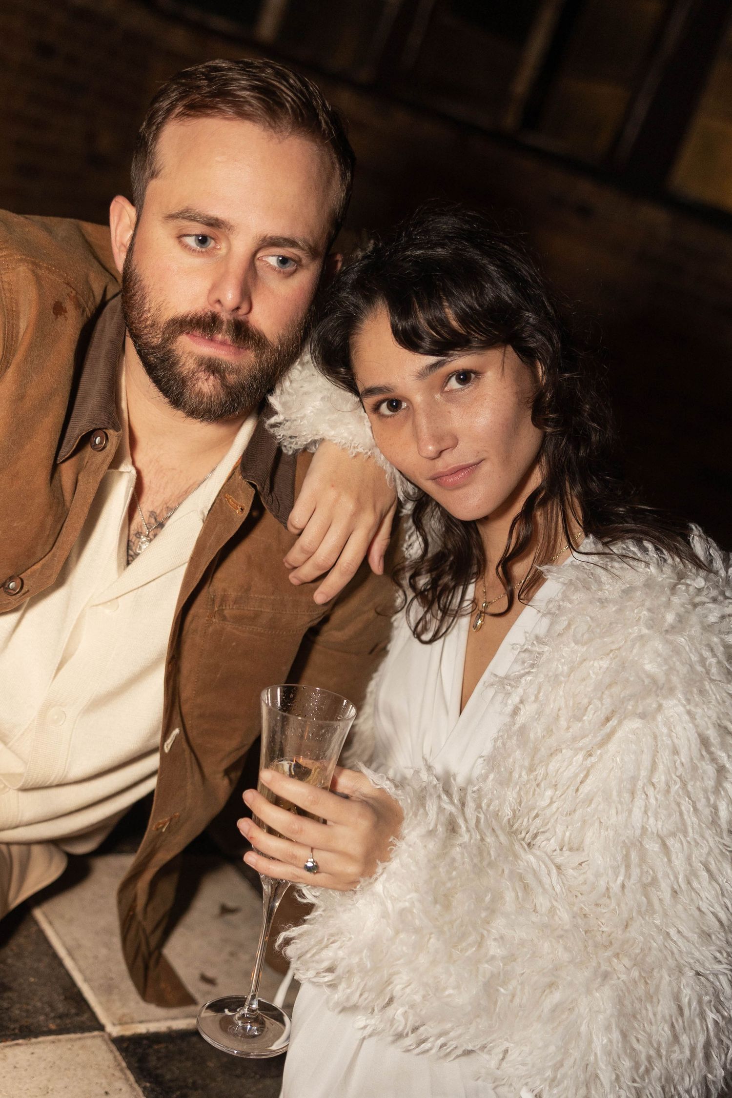 Stylish couple posing together at night, woman in white feathered jacket holding champagne flute, man in brown jacket.
