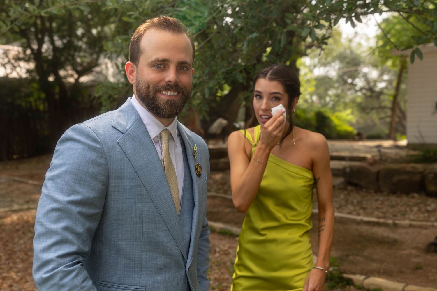 Groom in light blue suit smiles as bridesmaid in yellow dress wipes tears at outdoor wedding.