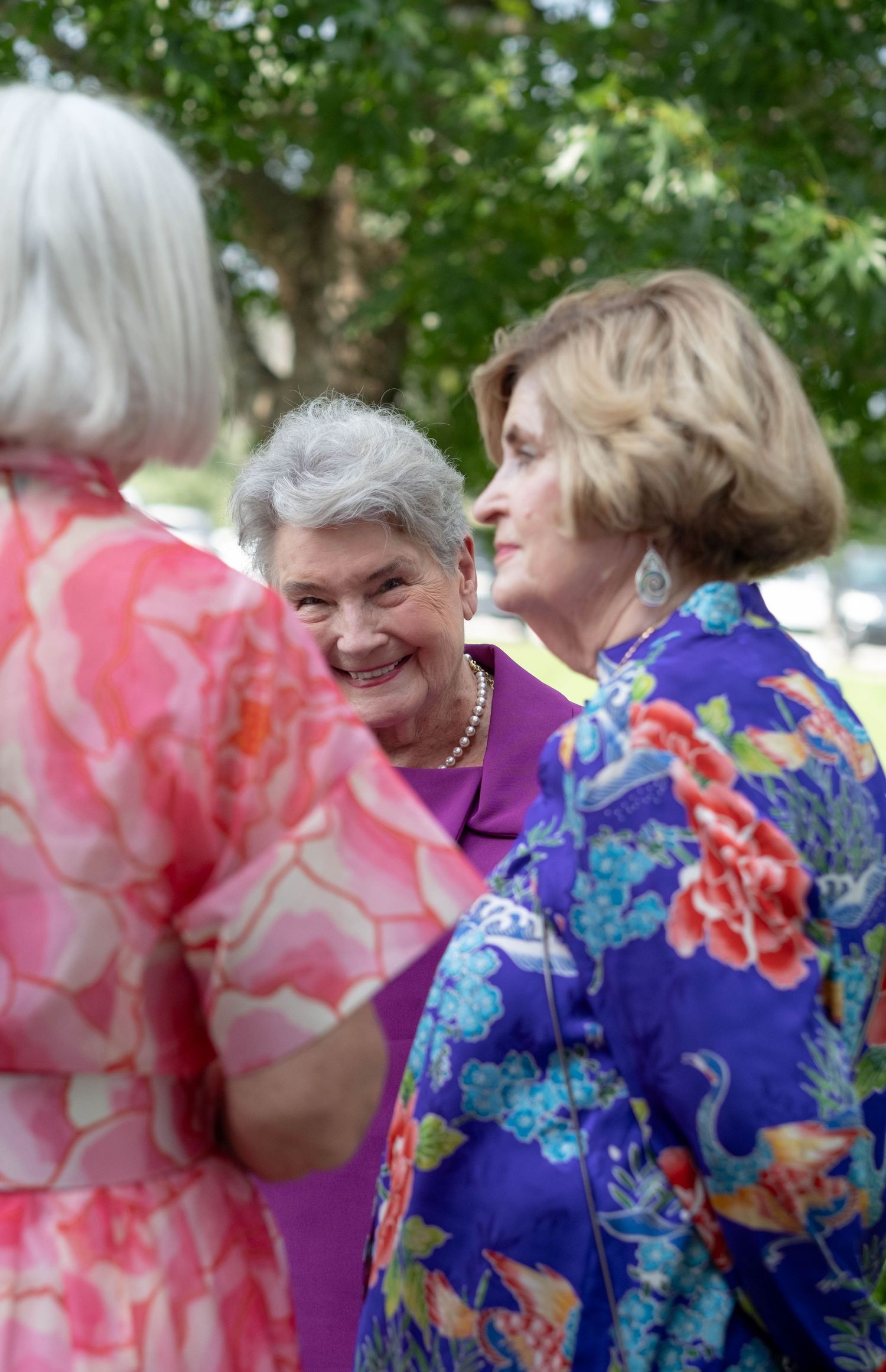 Three older women in floral dresses smiling and conversing outdoors under green trees.