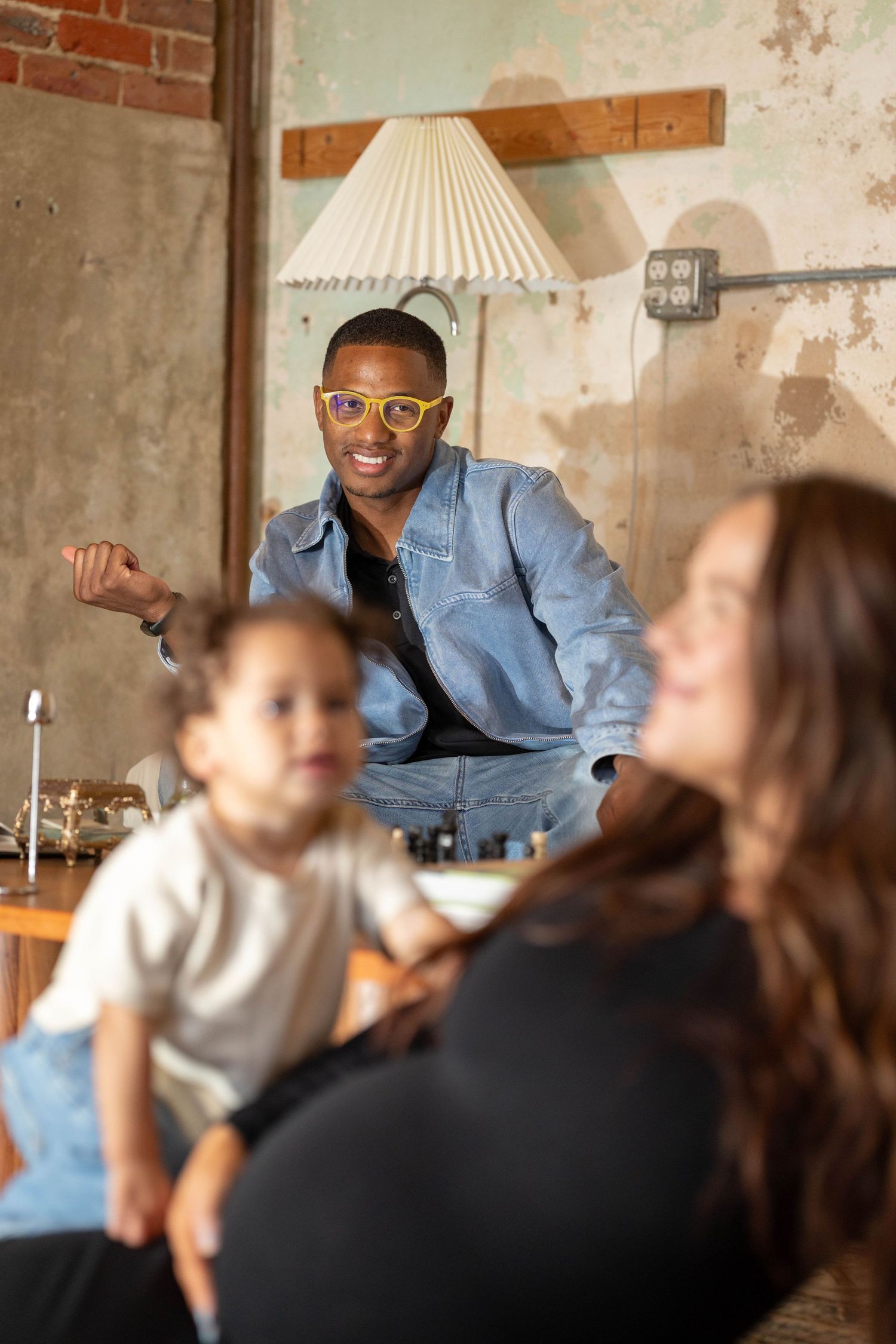 Man in denim jacket and yellow glasses smiling at camera with family nearby.