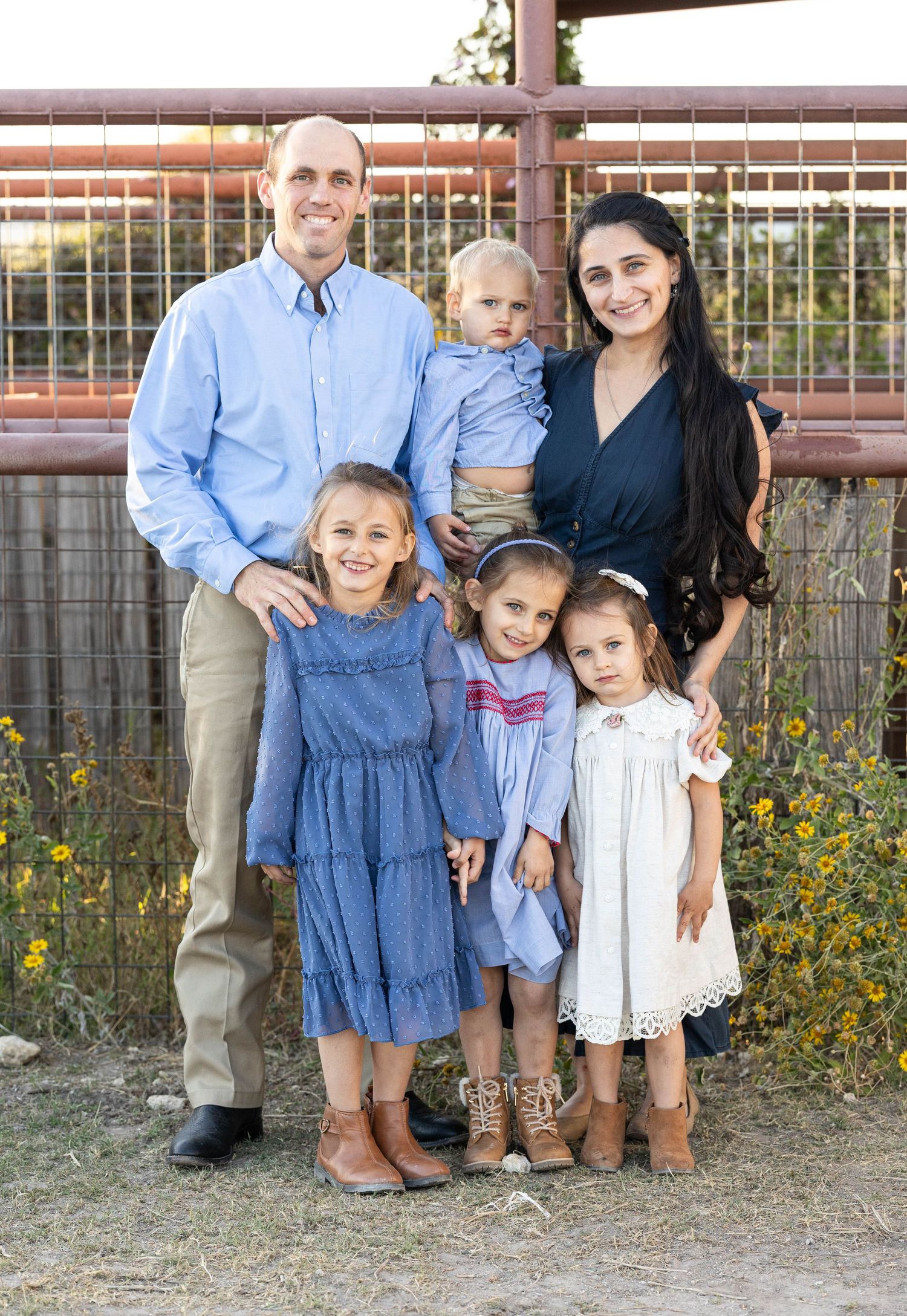 Family of five smiling outdoors near rustic metal fence, father and mother with three daughters and a toddler son.