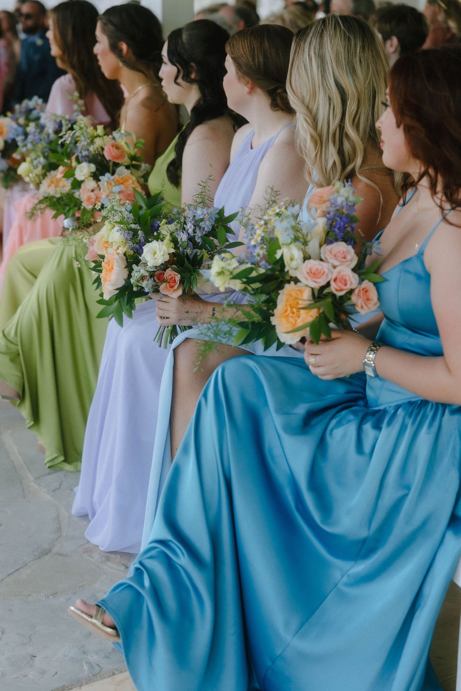 Bridesmaids seated in colorful mismatched satin gowns holding floral bouquets with peach roses and blue flowers at a wedding ceremony.