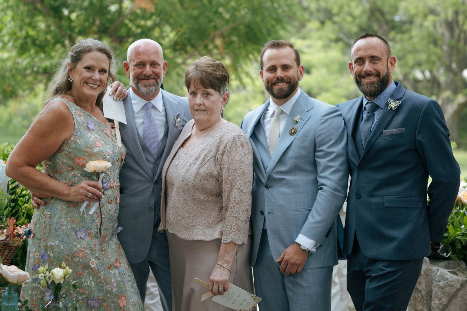 Group of five adults in formal attire smiling together outdoors at a wedding celebration.