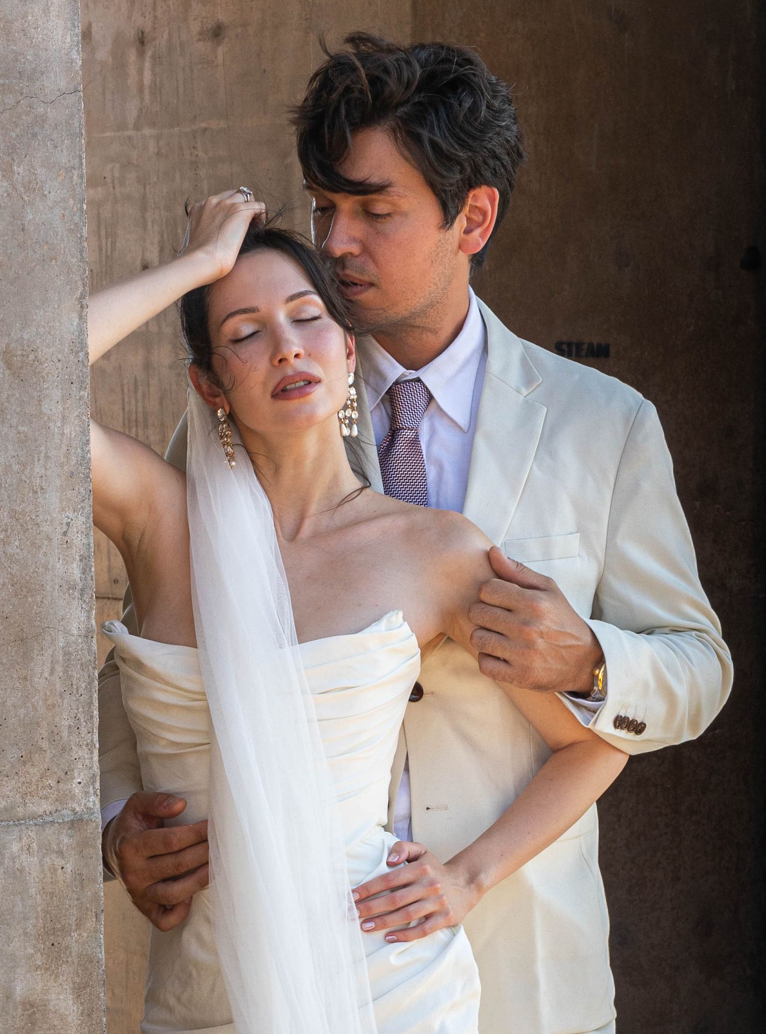 Bride in strapless gown and groom in cream suit pose intimately against a stone column outdoors.