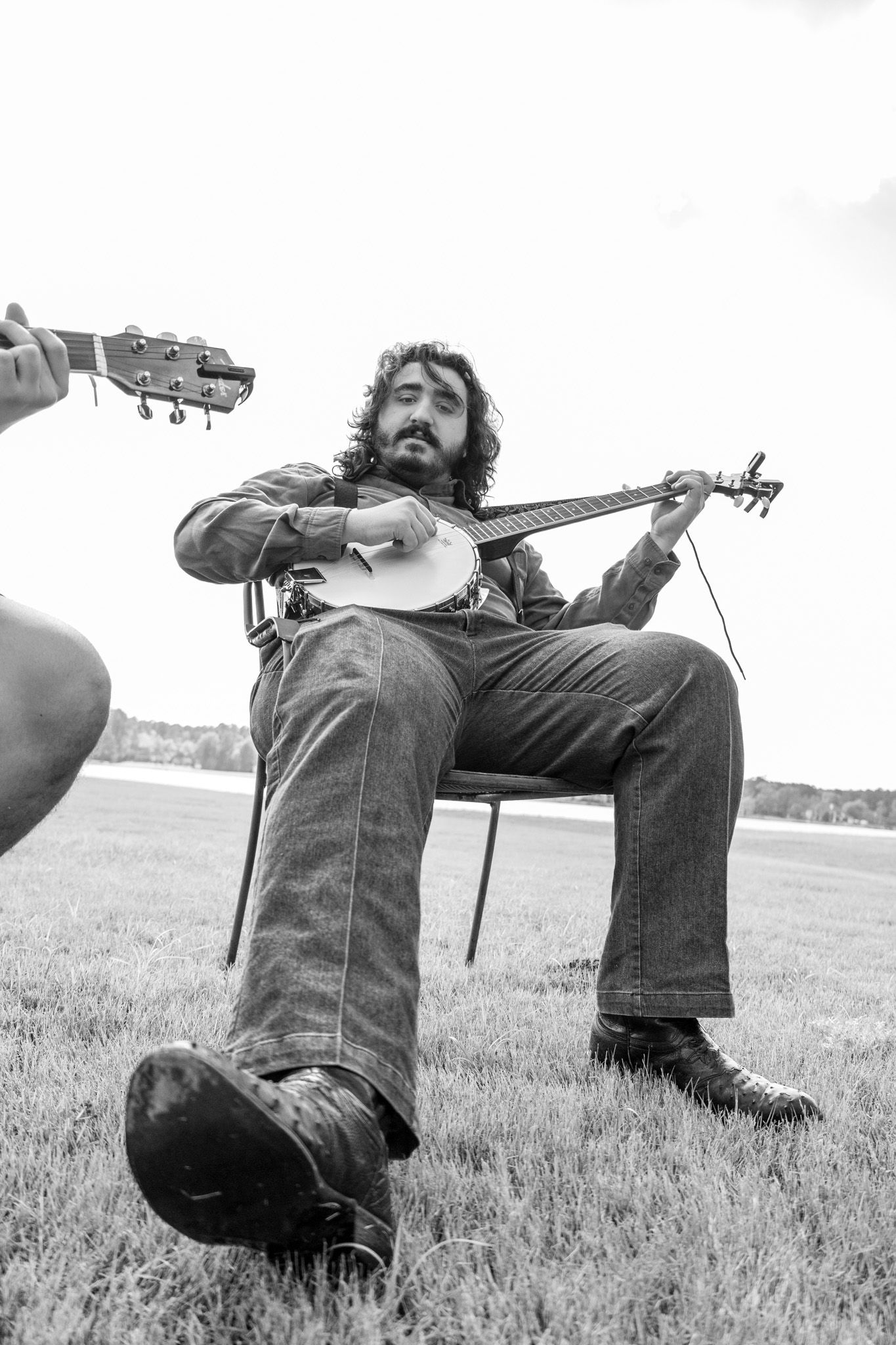 Bearded musician playing banjo while seated in chair on grass field in black and white photo.