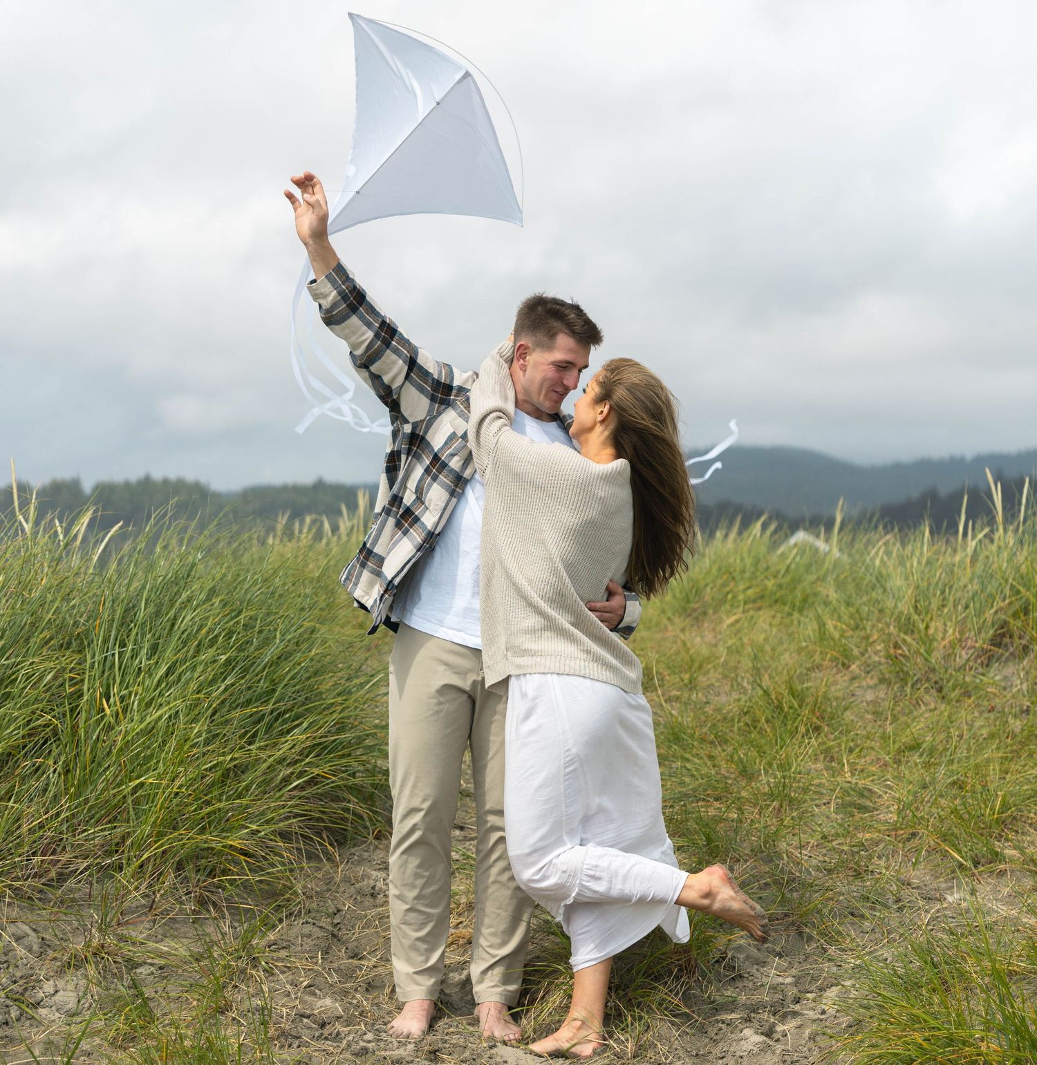 Couple kissing while holding white kite in windy coastal grass dunes on cloudy day.