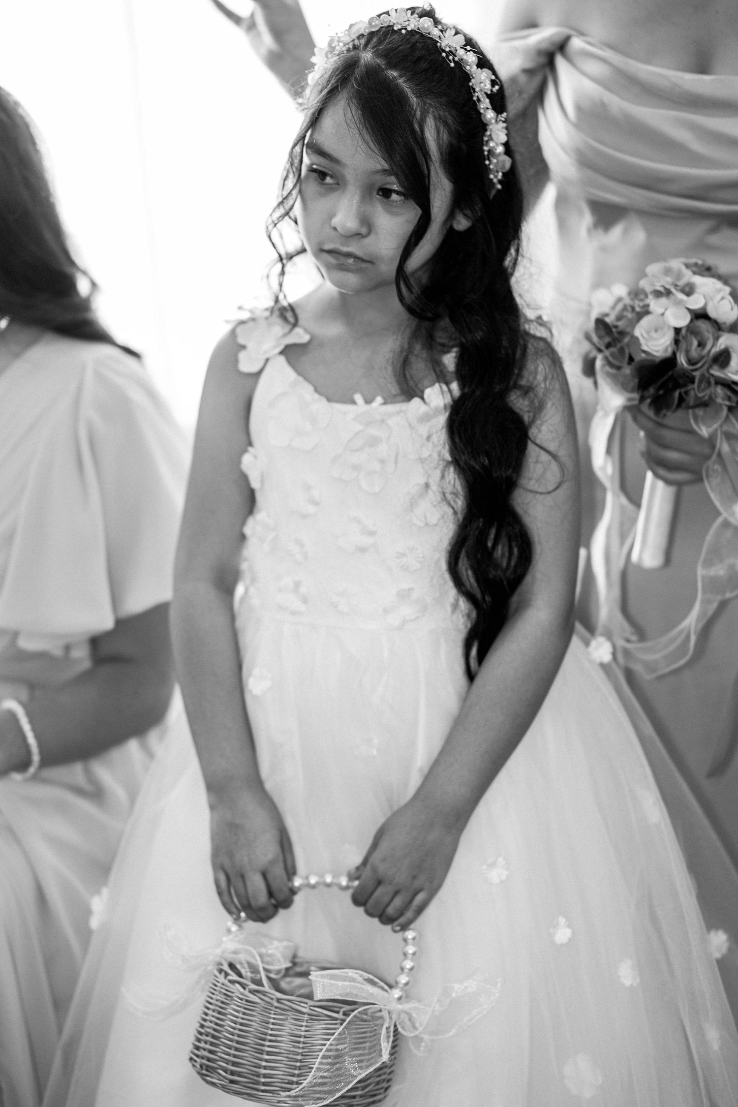Black and white portrait of flower girl in white dress holding basket at wedding ceremony.