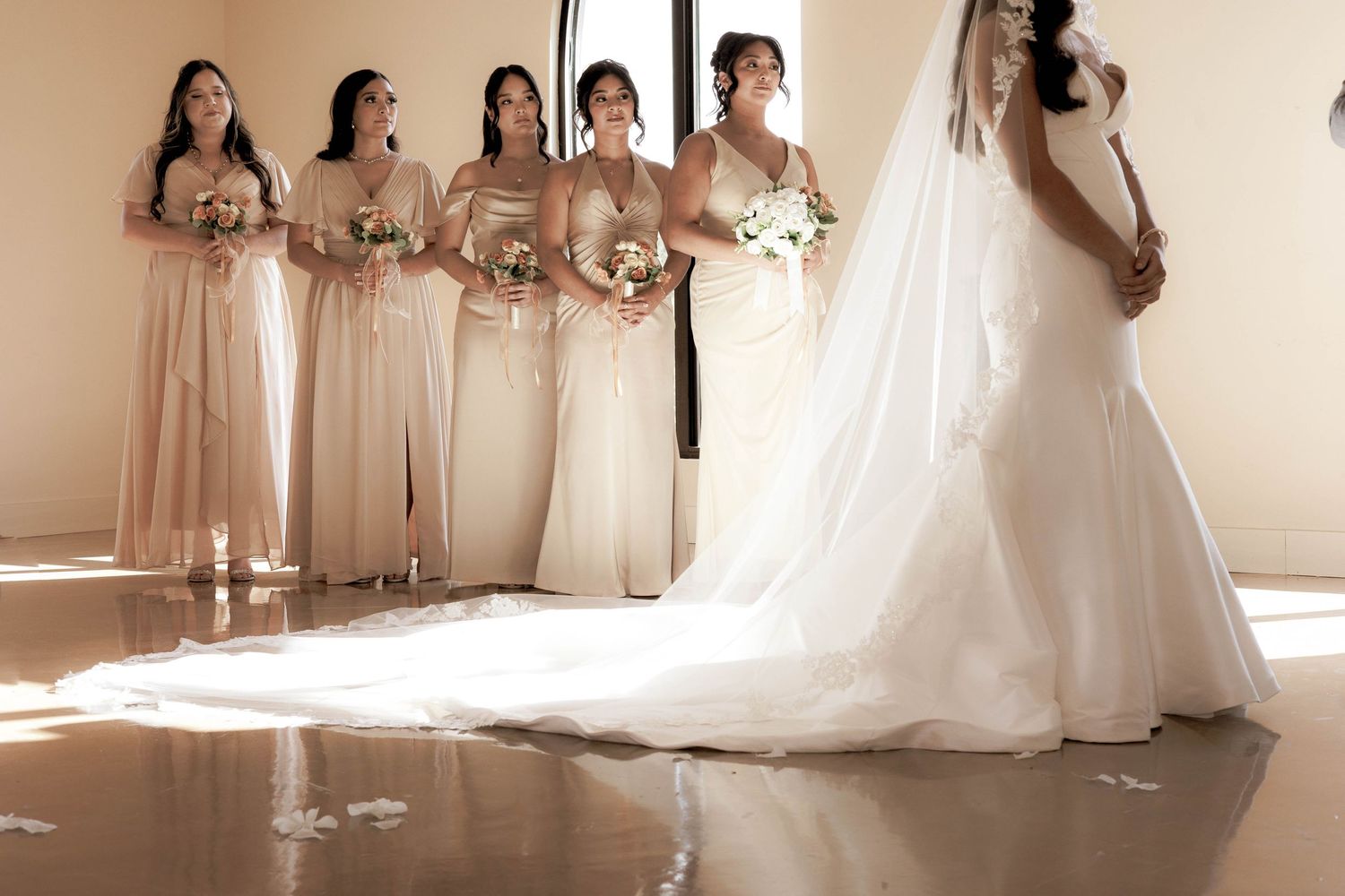 A bride in a white mermaid gown with a long veil stands before her bridesmaids in champagne dresses holding bouquets.