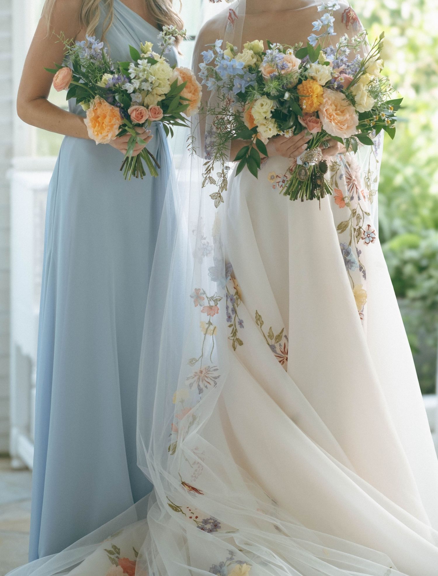 Bride in floral embroidered wedding gown and bridesmaid in blue dress holding pastel bouquets.
