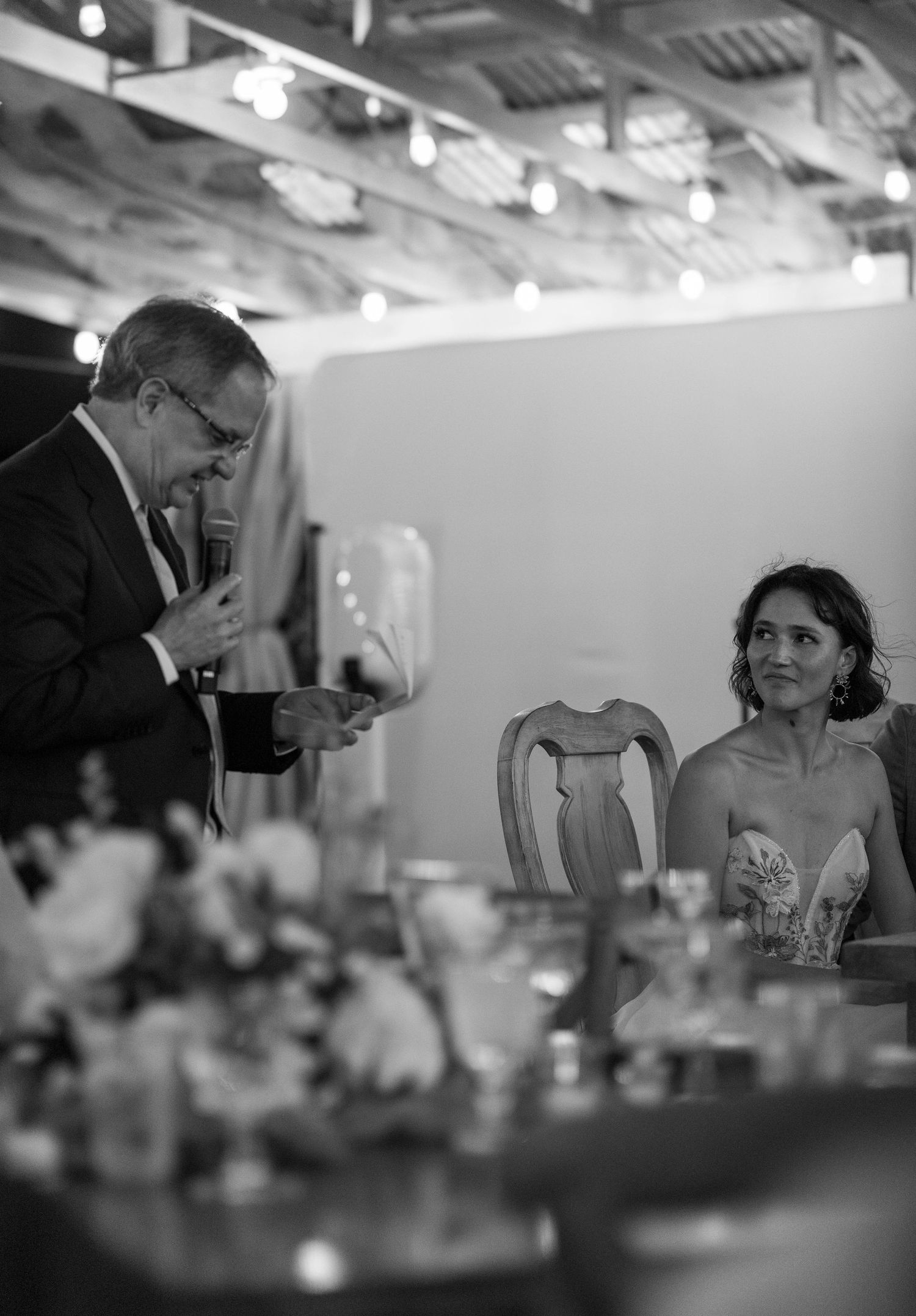 Black and white photo of father giving emotional speech while bride listens at reception.