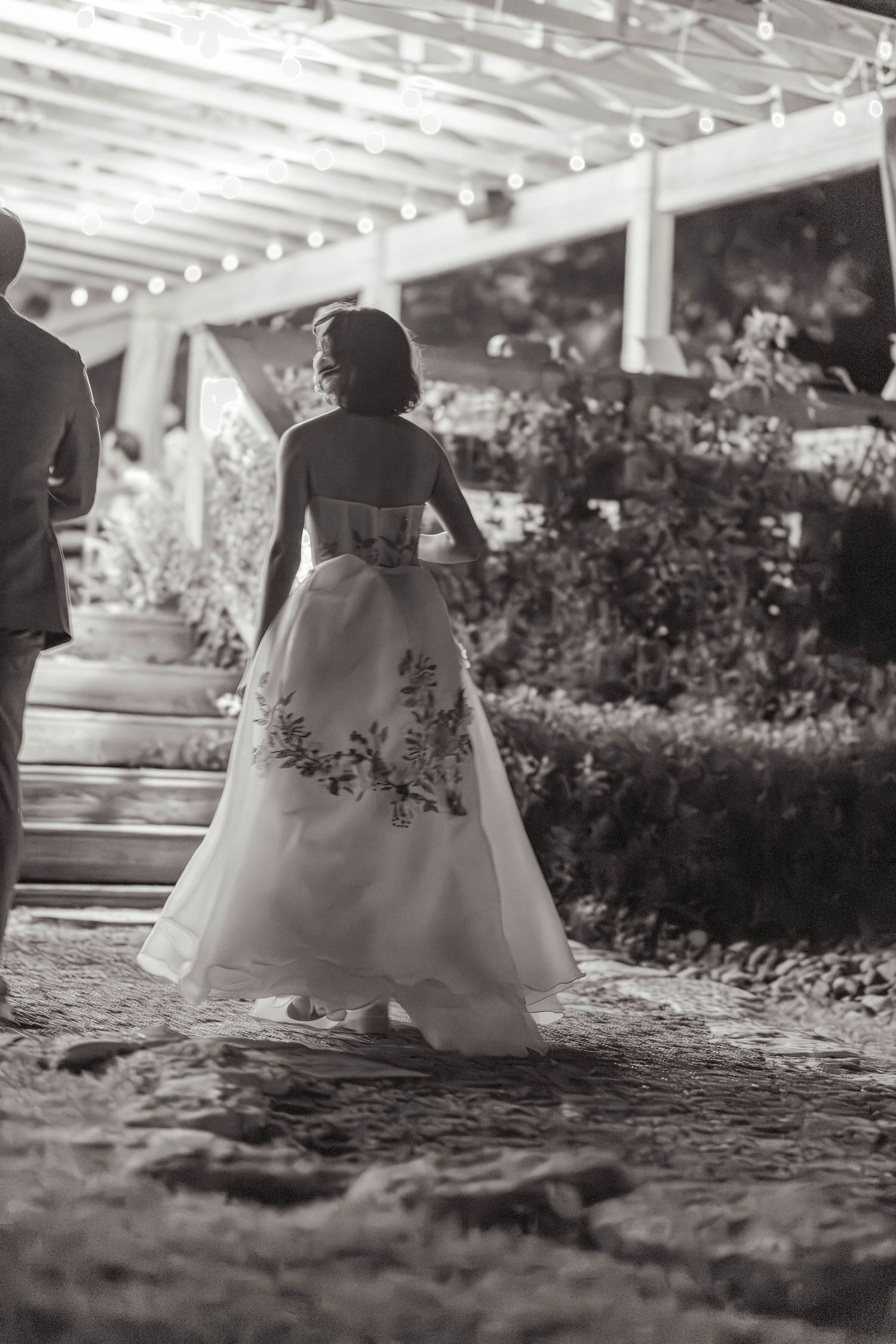 Black and white rear view of bride in floral embroidered gown walking toward lit reception venue.