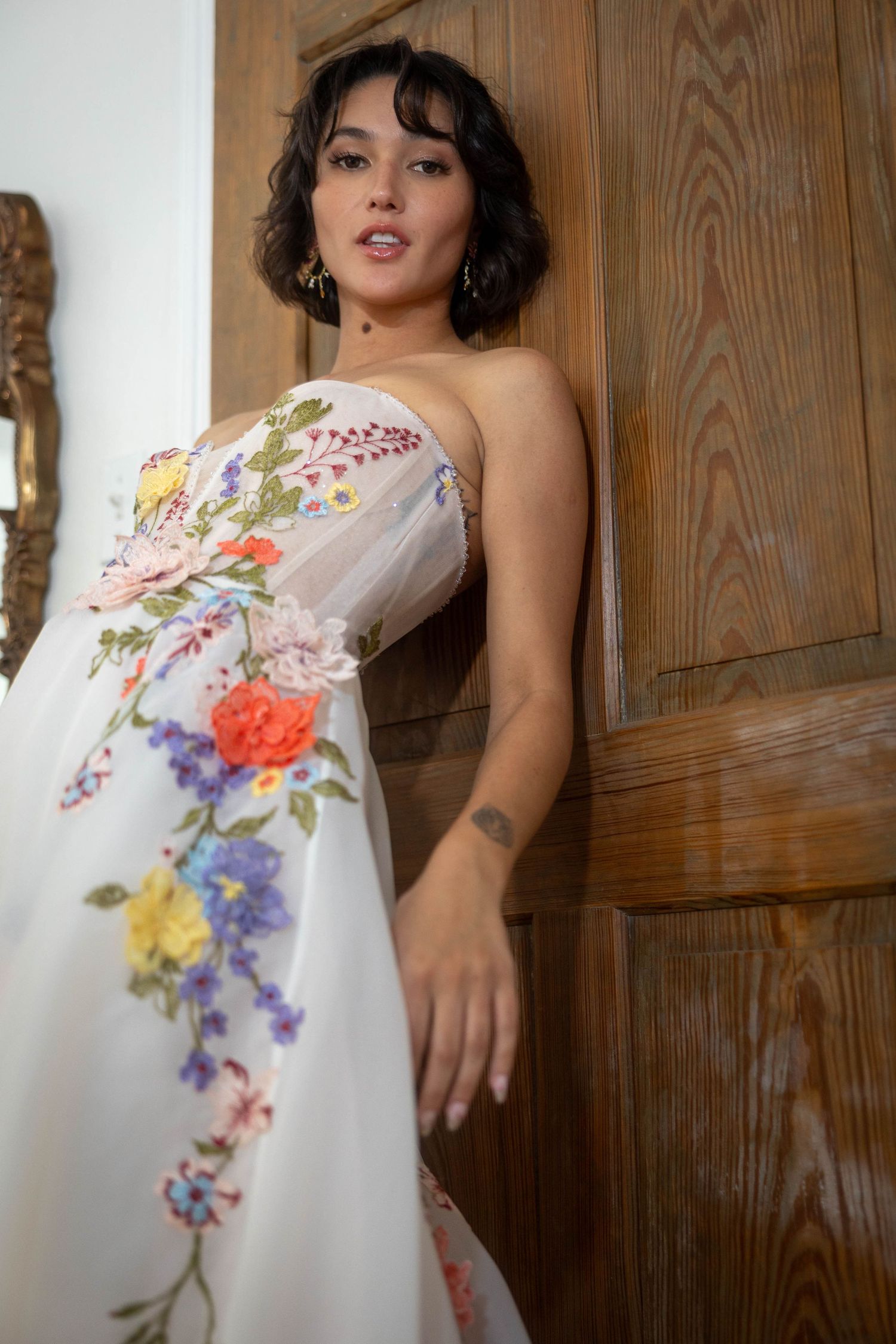 Close-up of bride in a strapless white gown with colorful 3D floral embroidery leaning against a wooden door.