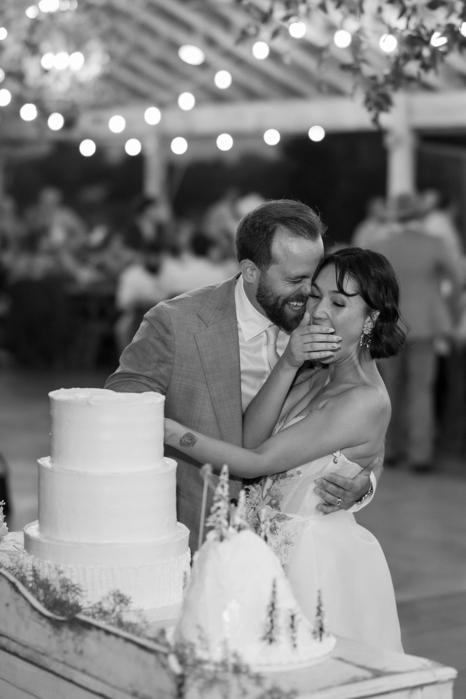 Black and white photo of a groom kissing his bride beside a three-tier wedding cake under string lights at reception.