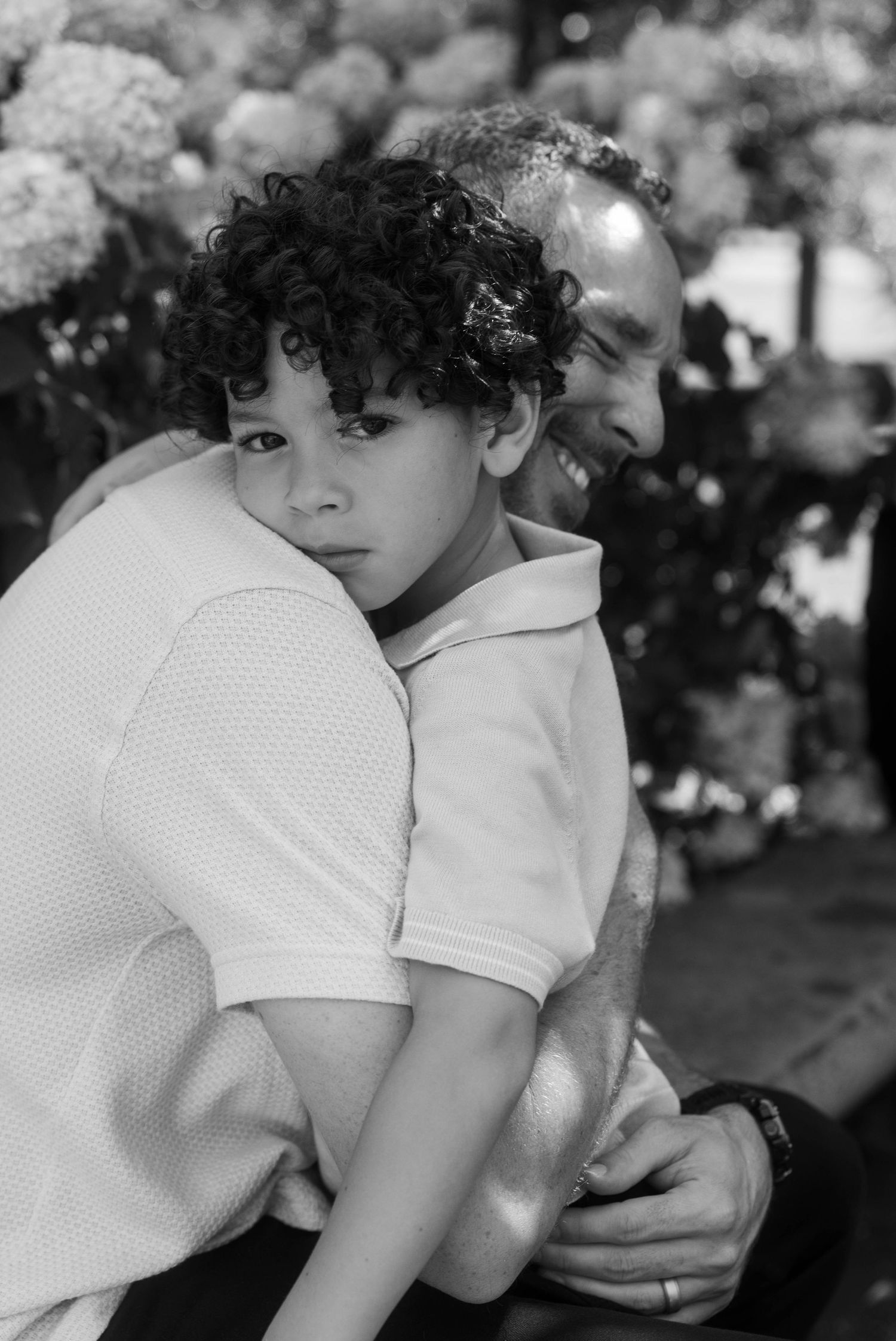 Black and white photo of a curly-haired boy hugging a smiling man outdoors near blooming flowers.
