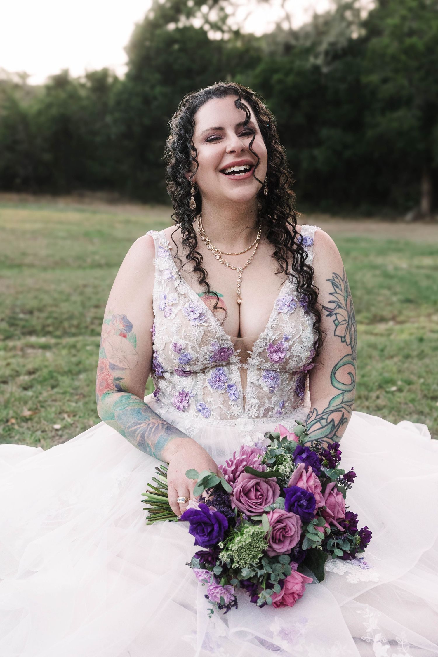 Smiling tattooed bride in floral wedding dress sits outdoors holding vibrant purple and pink flower bouquet.