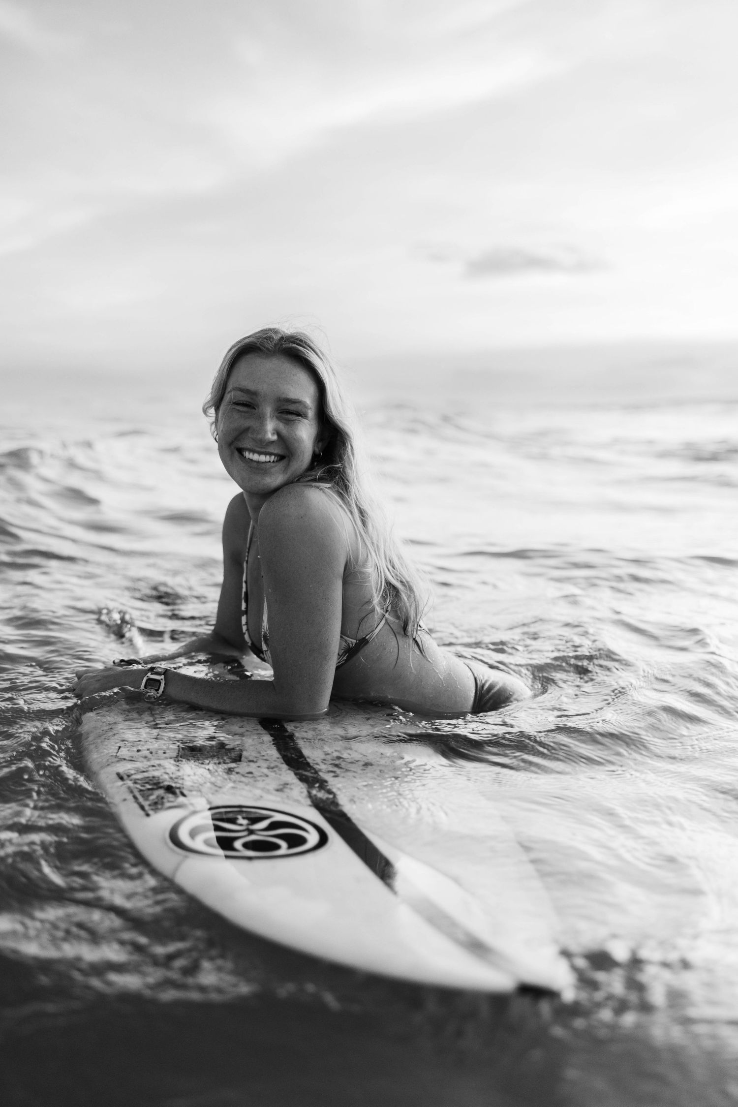 Black and white photo of smiling woman lying on surfboard in ocean waves.