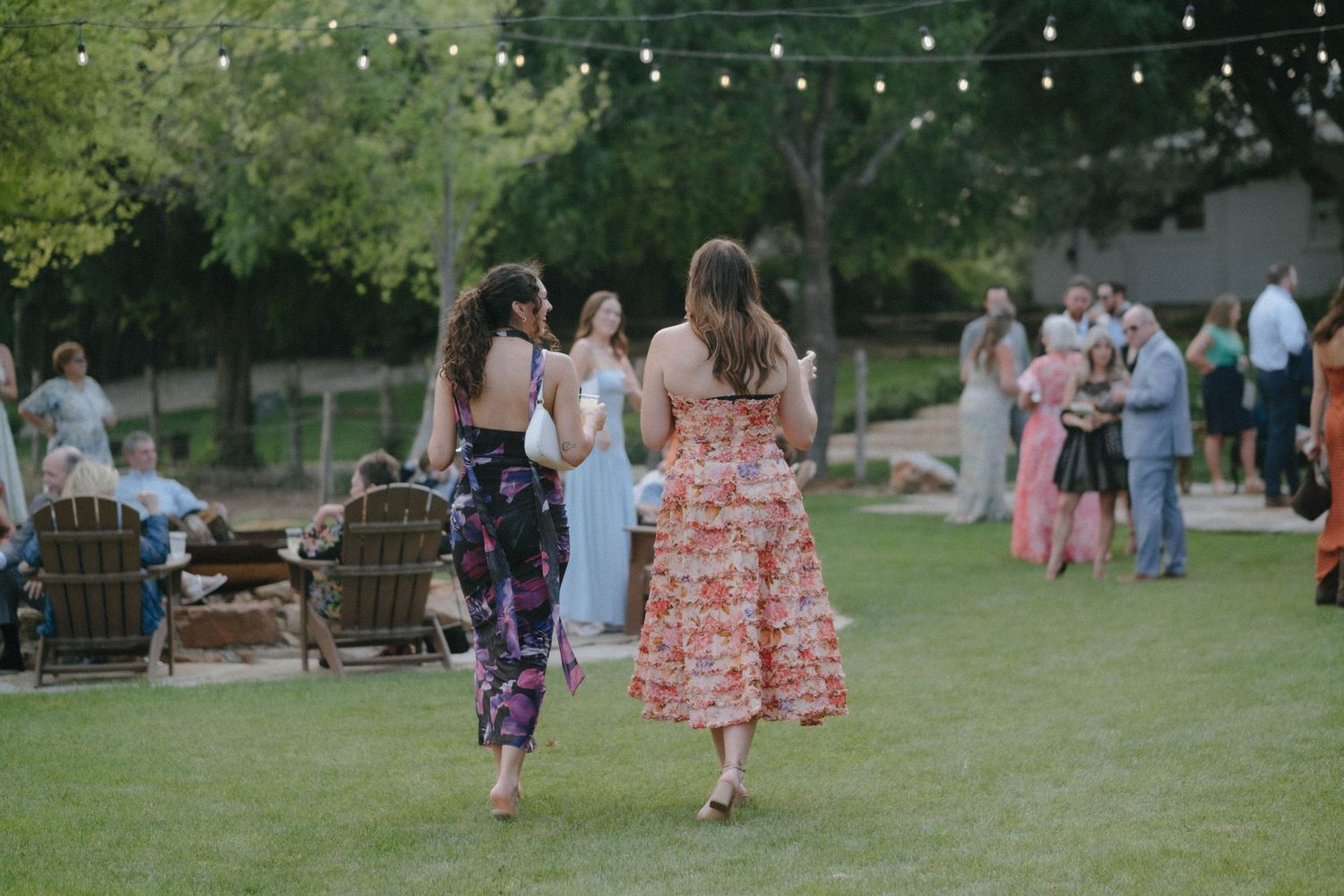 Two guests in colorful dresses walk across a green lawn toward wedding cocktail hour festivities.