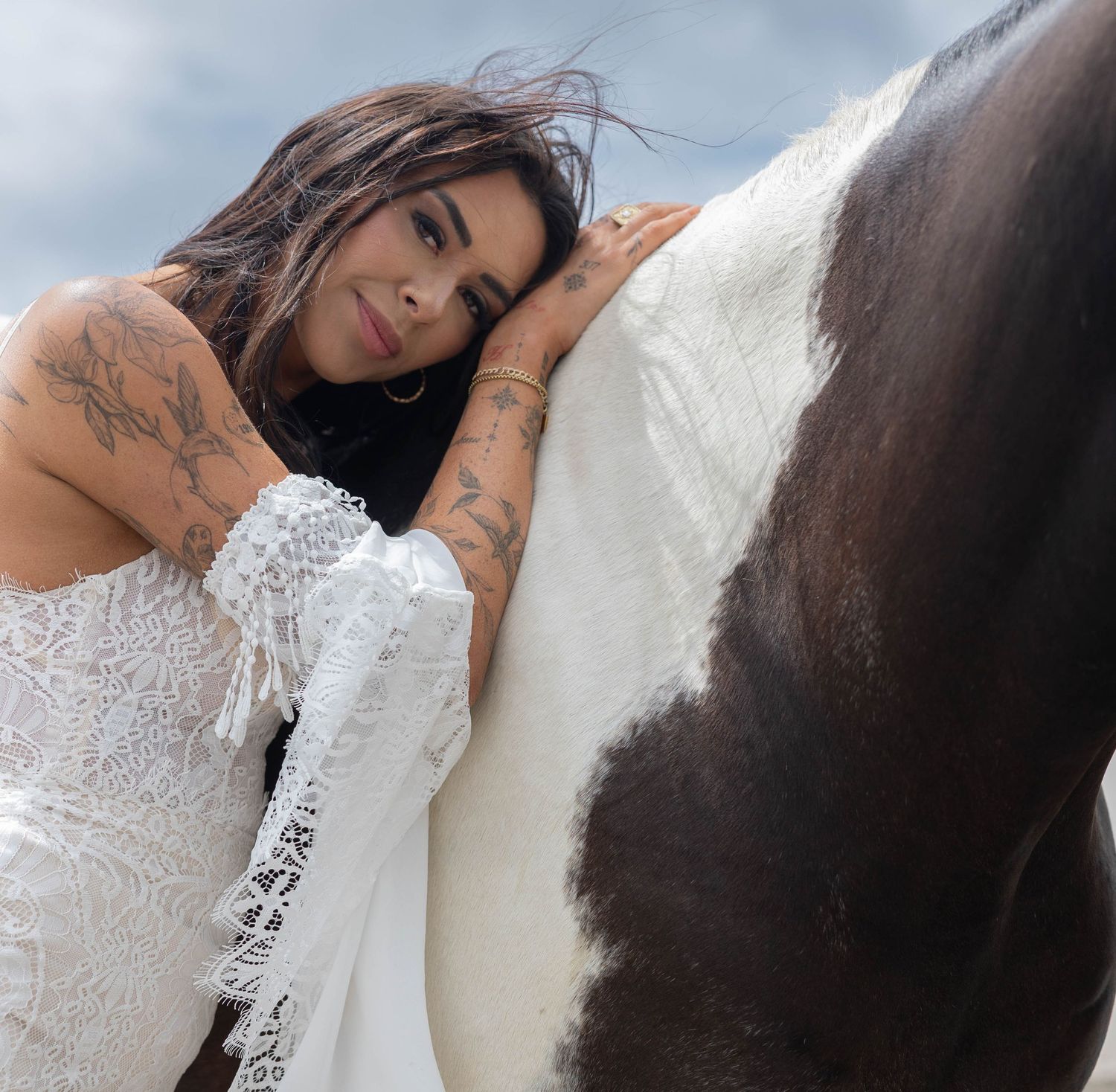Tattooed bride in white lace dress leaning against a paint horse outdoors.