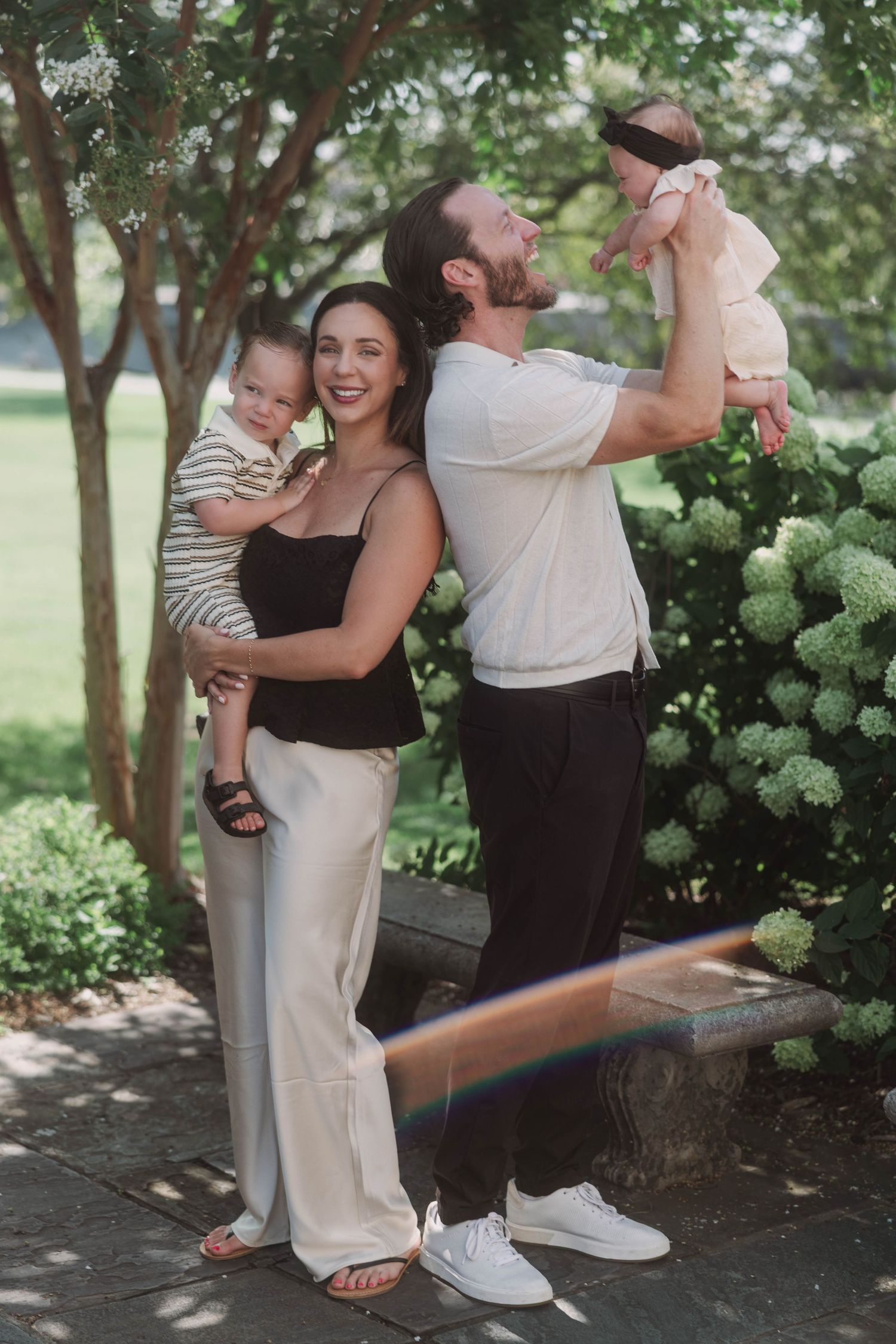 Happy family posing outdoors with two babies, surrounded by lush green trees and white hydrangea bushes.