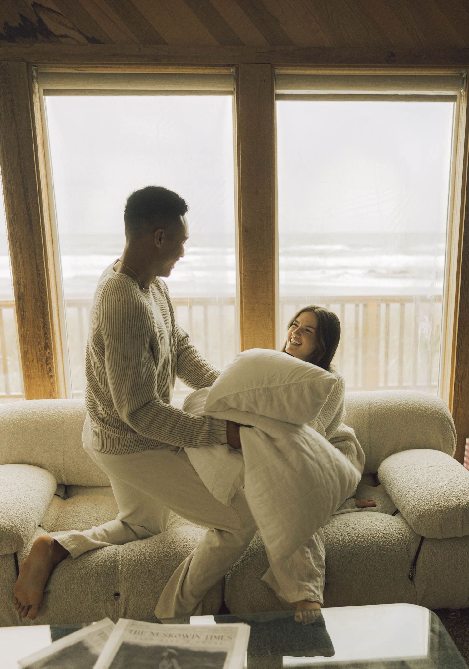 A playful couple laughs together on a sofa, with one holding a large pillow, ocean views in background.