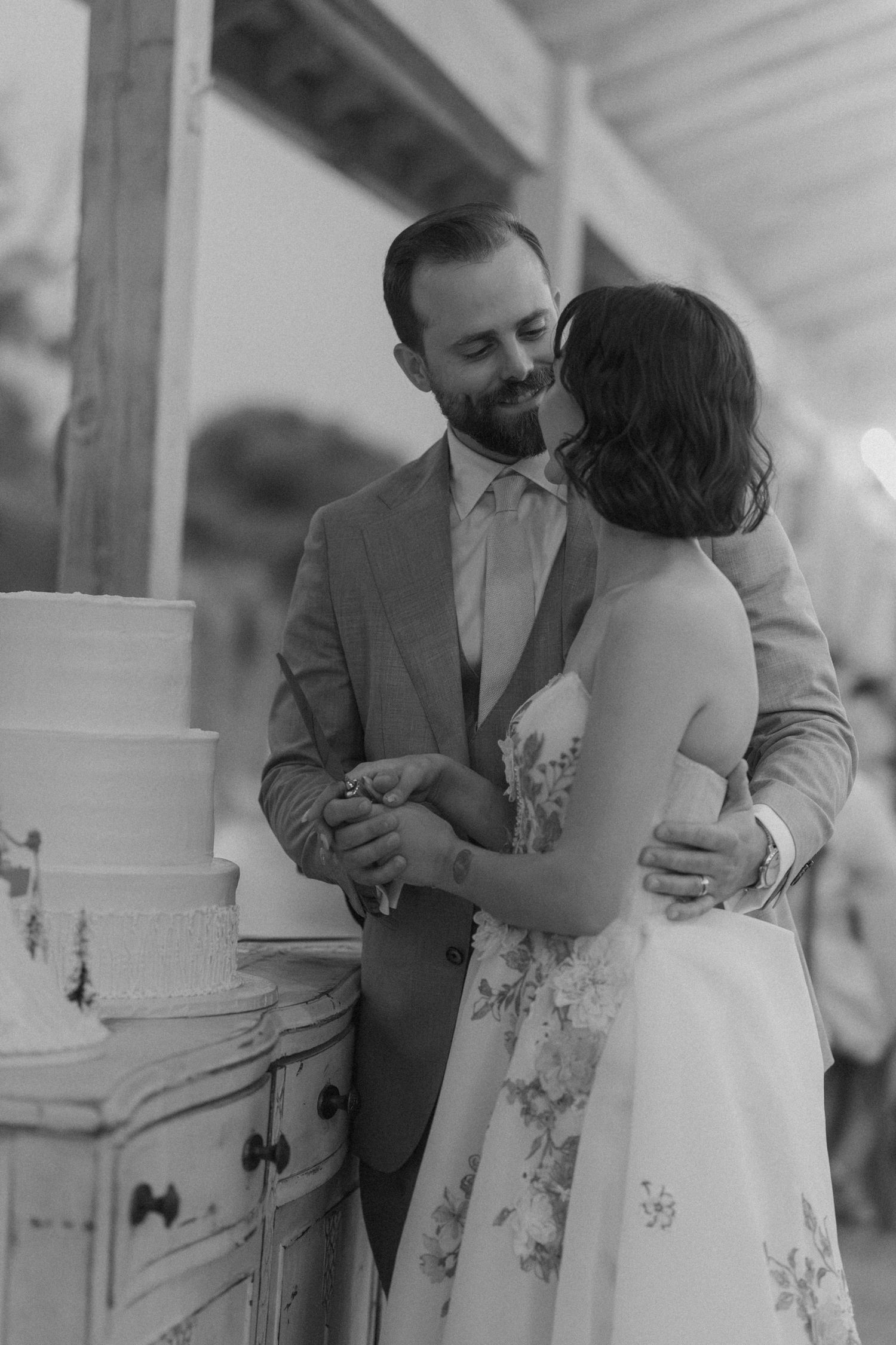 Black and white photo of groom kissing bride as they cut their wedding cake together.