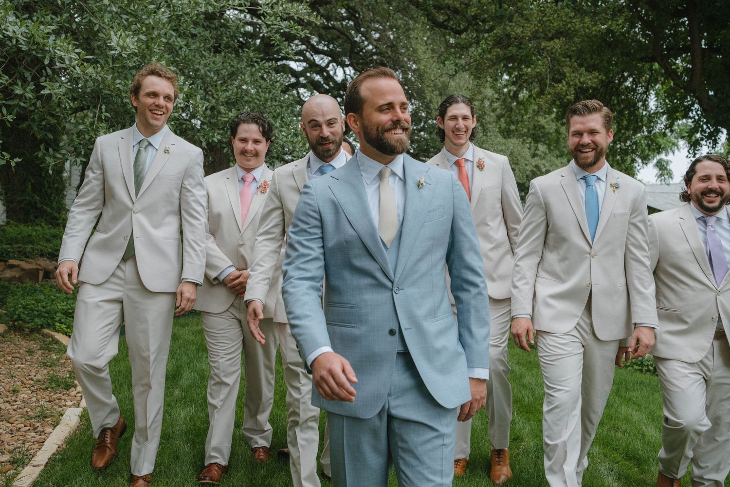 Groom in light blue suit leads groomsmen in tan suits walking outdoors at wedding.