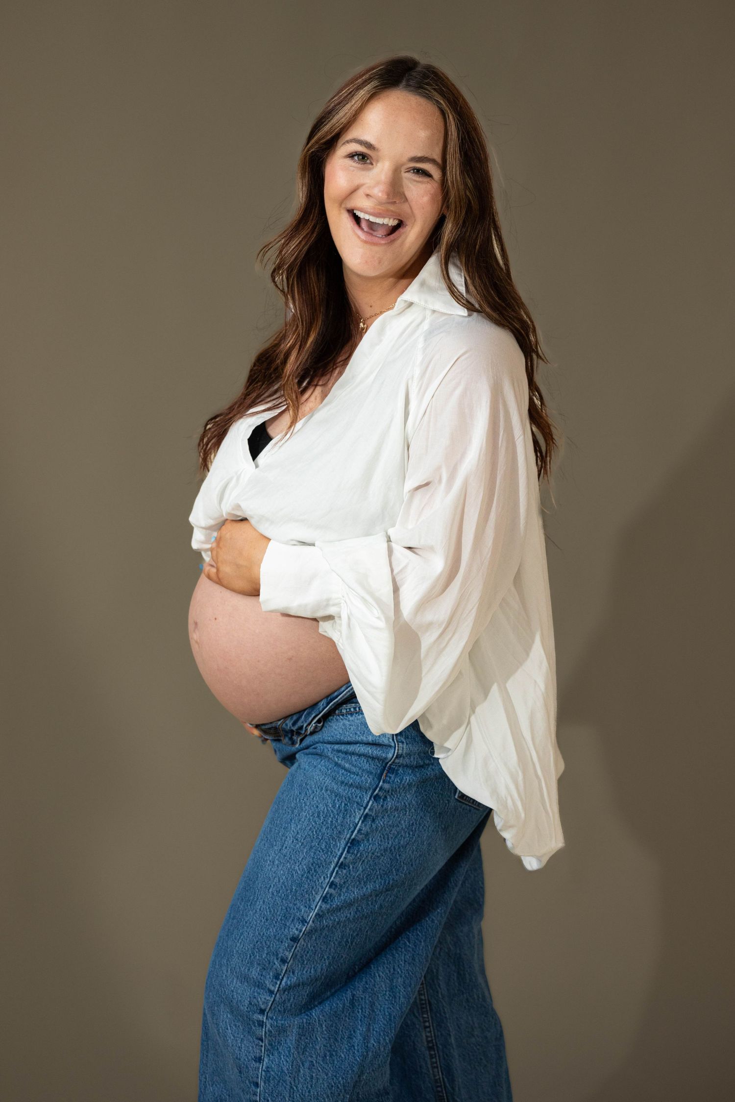 Laughing pregnant woman in white shirt and jeans showing bare bump on gray backdrop.