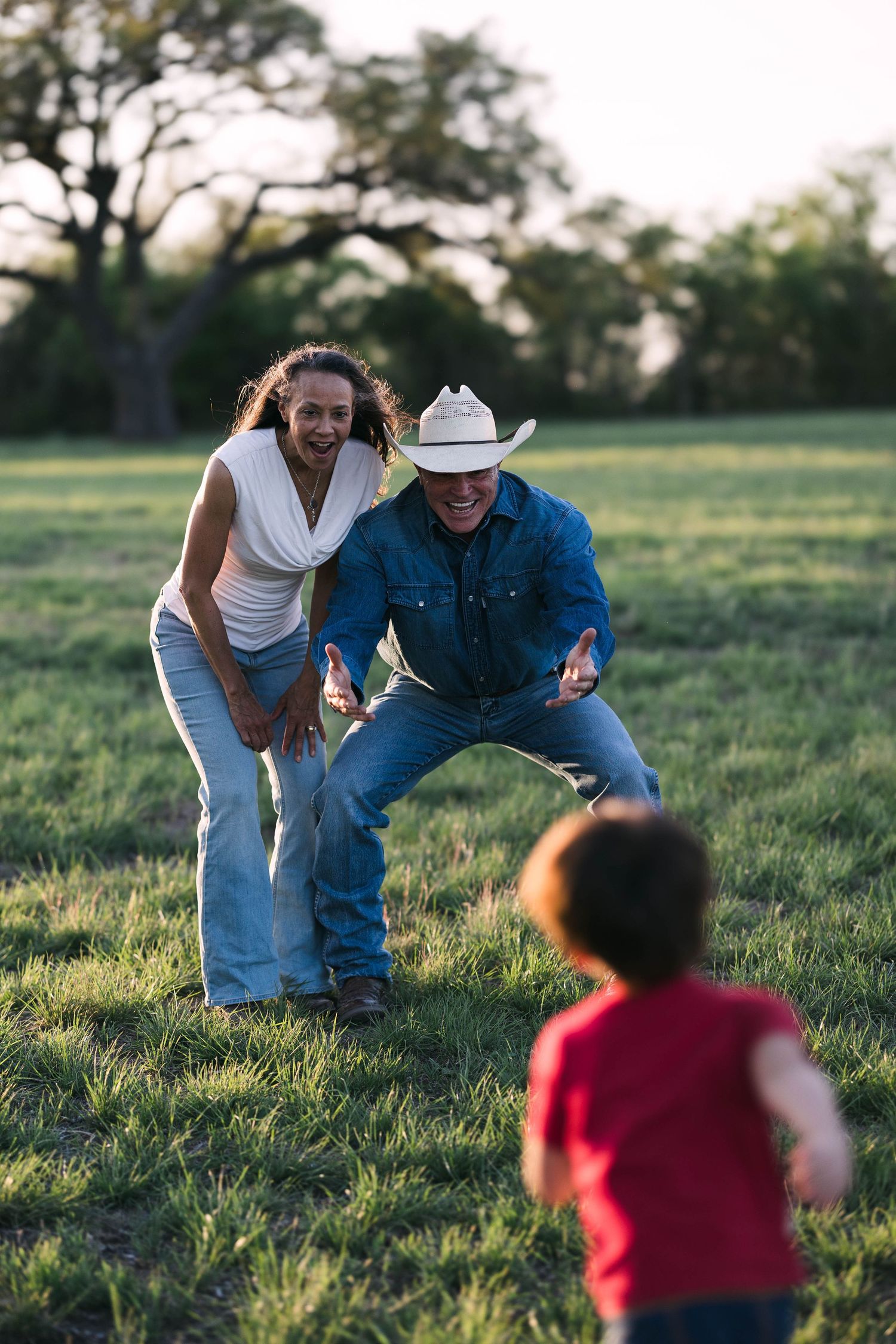 Laughing couple in a green field crouching down as a young child runs toward them at dusk.