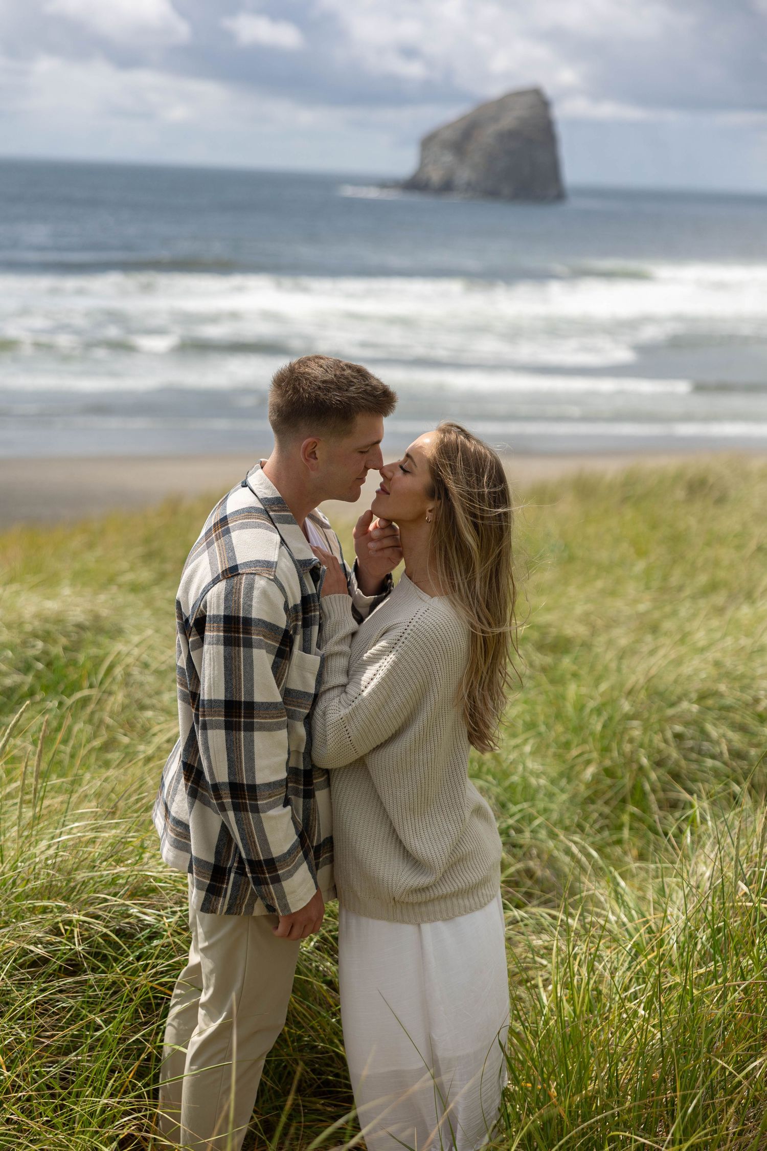 Couple sharing a romantic moment in tall coastal grass with ocean and sea stack visible in the background.