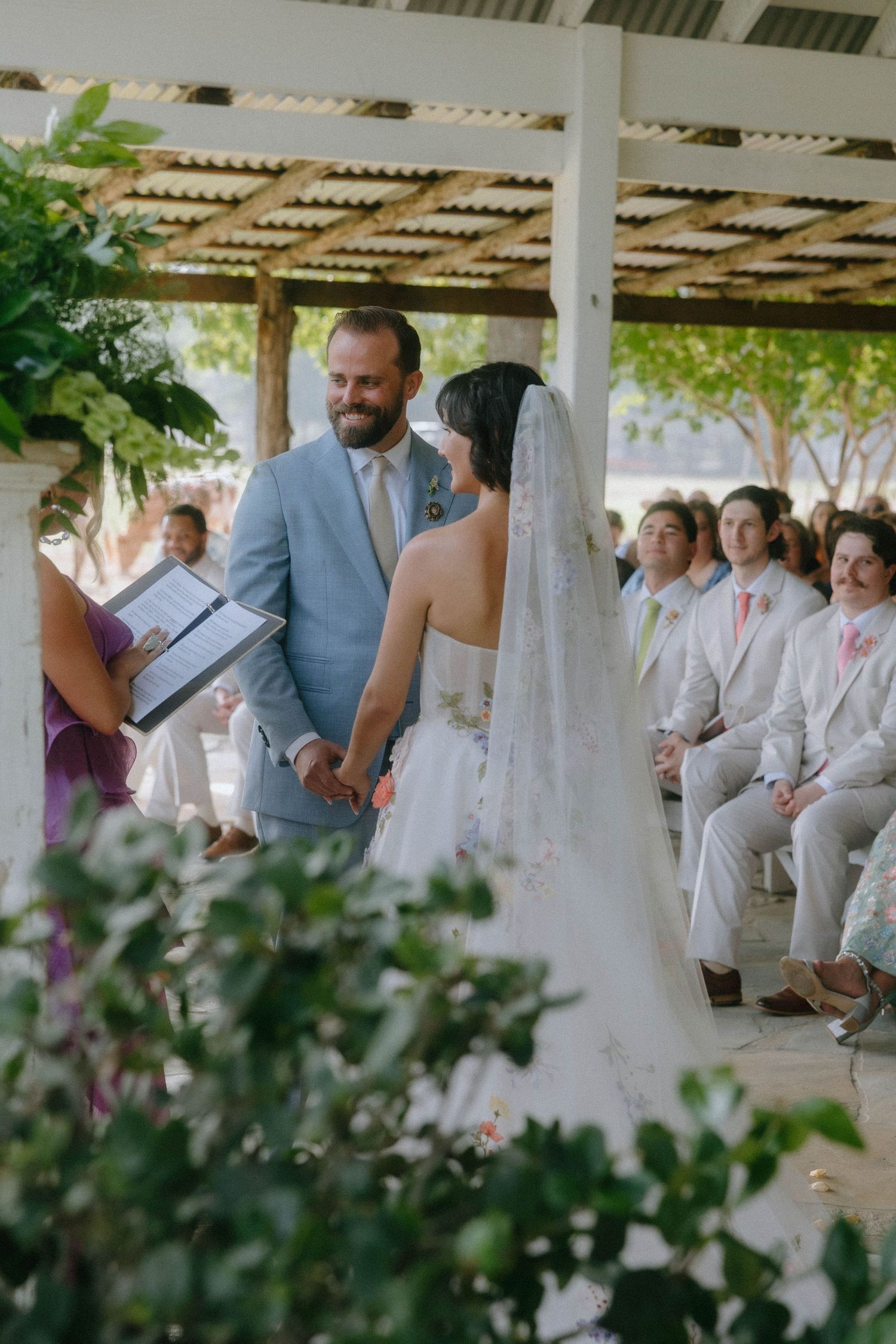 Bride and groom exchange vows under a rustic pergola during an outdoor wedding ceremony.