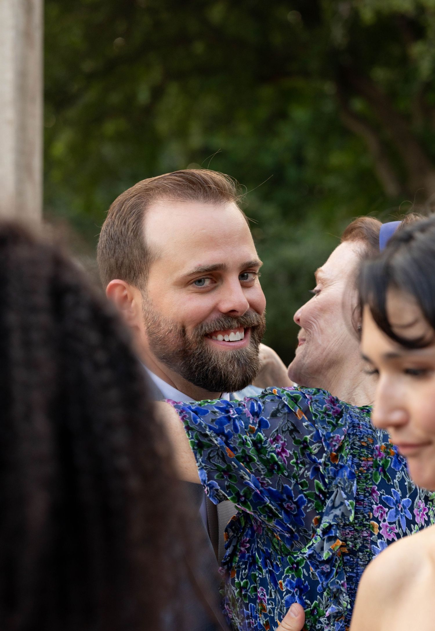 Bearded groom smiling warmly at wedding guest in blue floral dress outdoors.