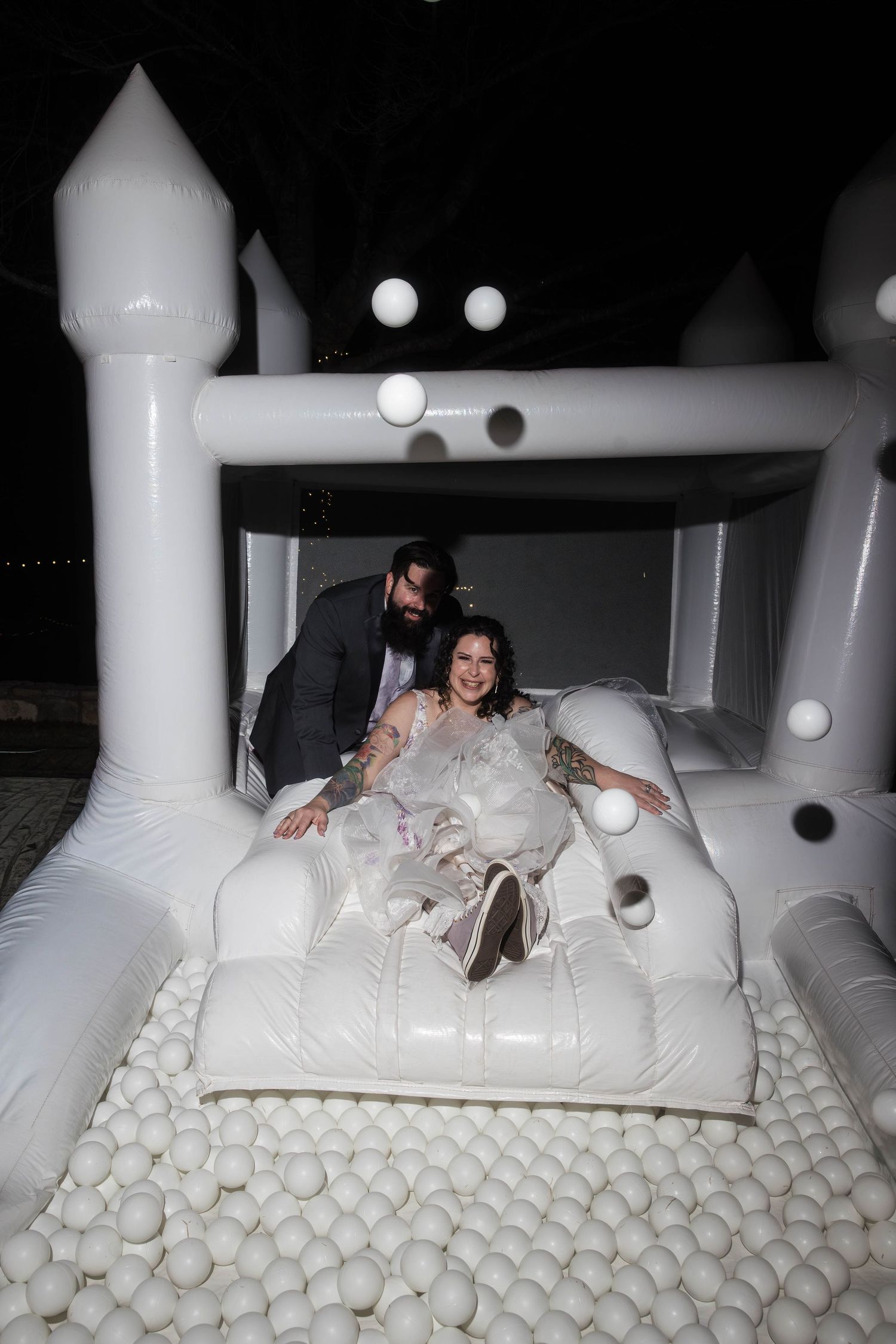Laughing bride and bearded groom play in a white inflatable bounce house ball pit at their wedding reception.