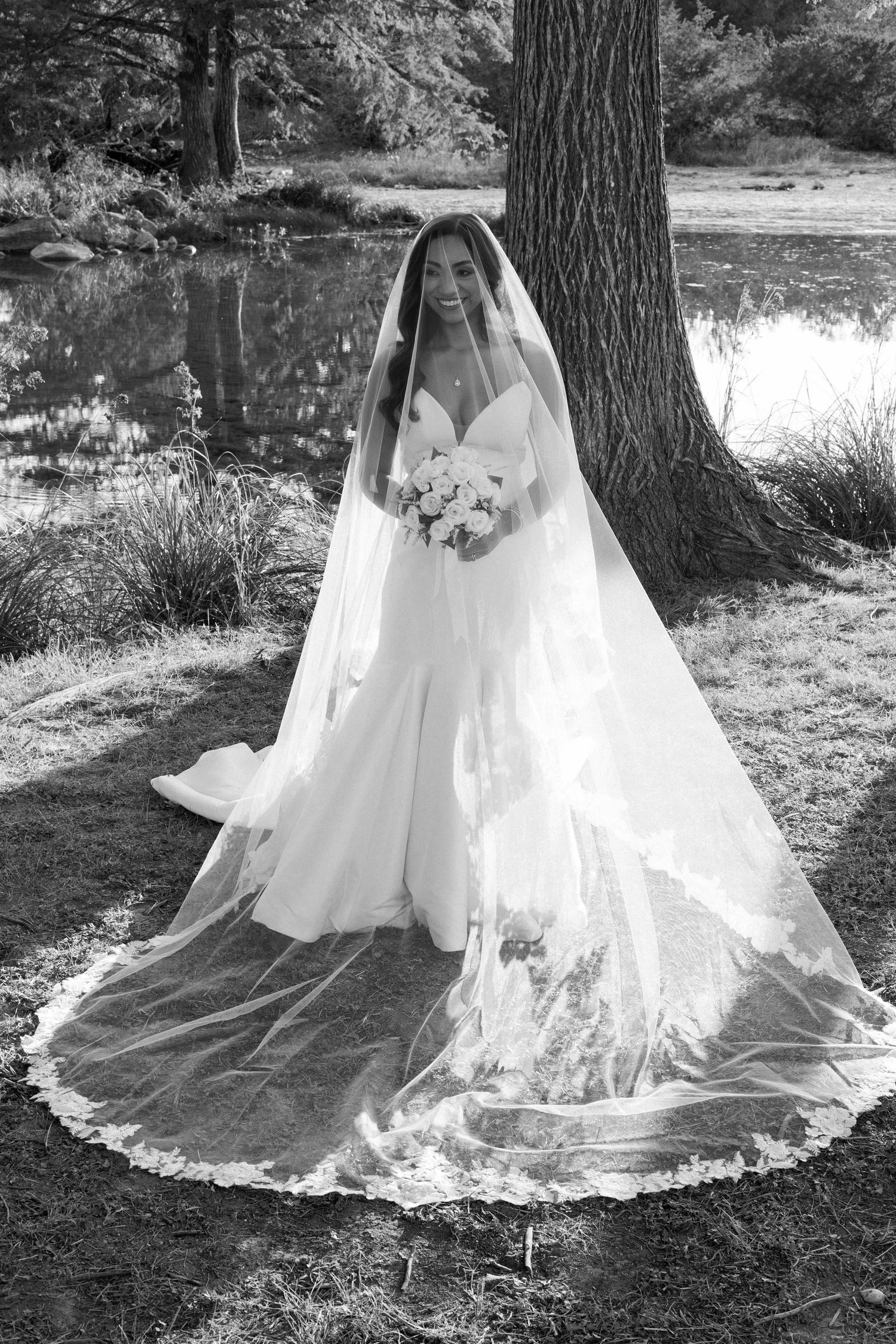 Black and white photo of a bride in a flowing wedding gown and long lace veil holding a bouquet by a lakeside.