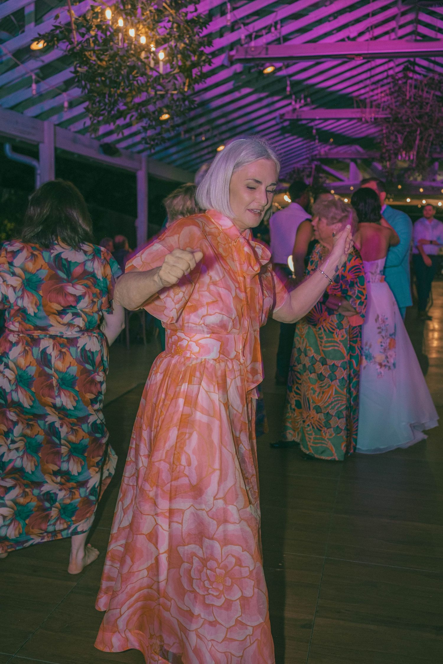 Woman in orange floral maxi dress dances joyfully under string lights and chandelier at wedding reception.