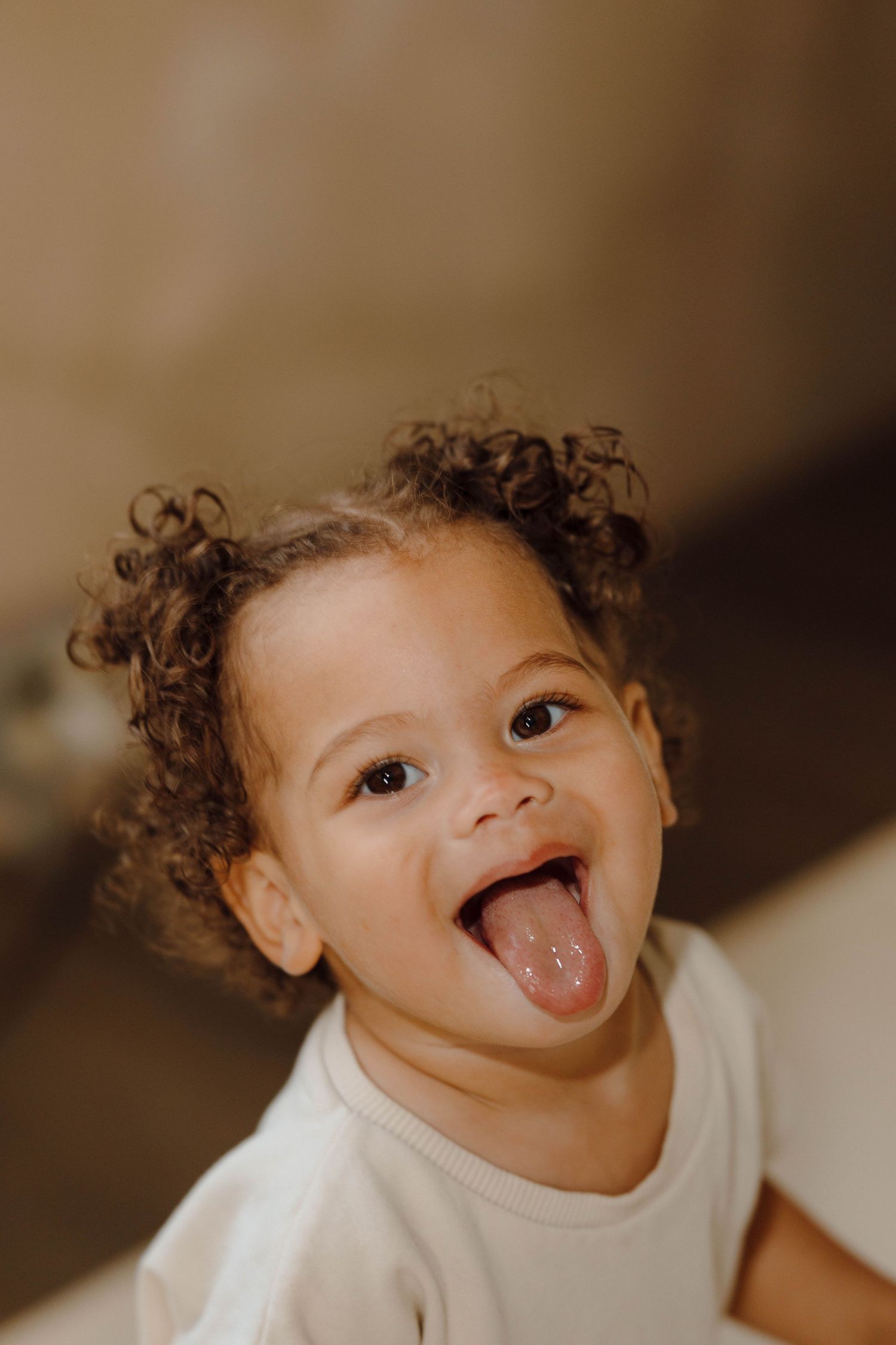 Adorable curly-haired toddler in white onesie sticks tongue out playfully in close-up portrait photo.