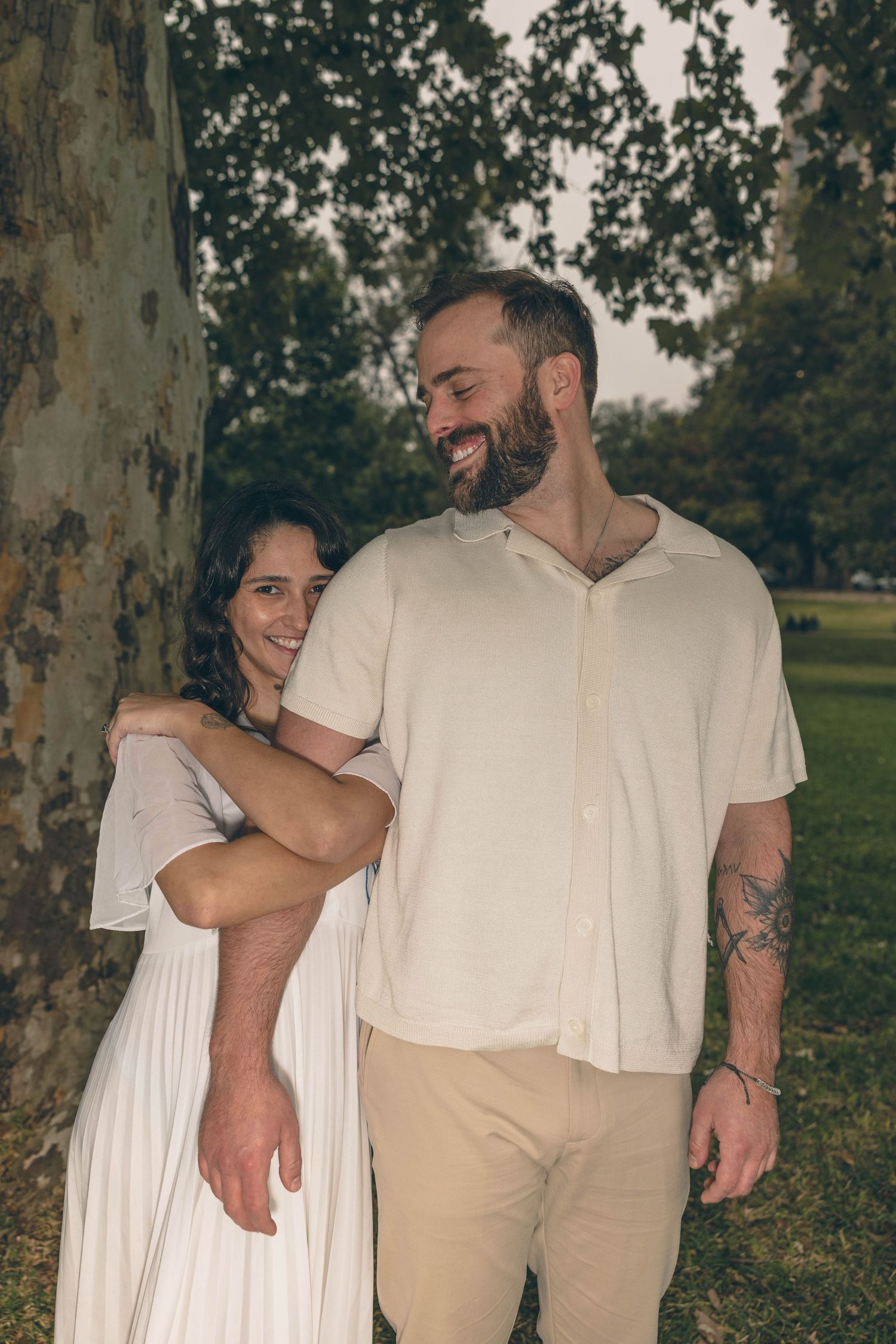 Couple leaning against large tree in park, woman in white dress smiling, man in cream shirt with tattoo.