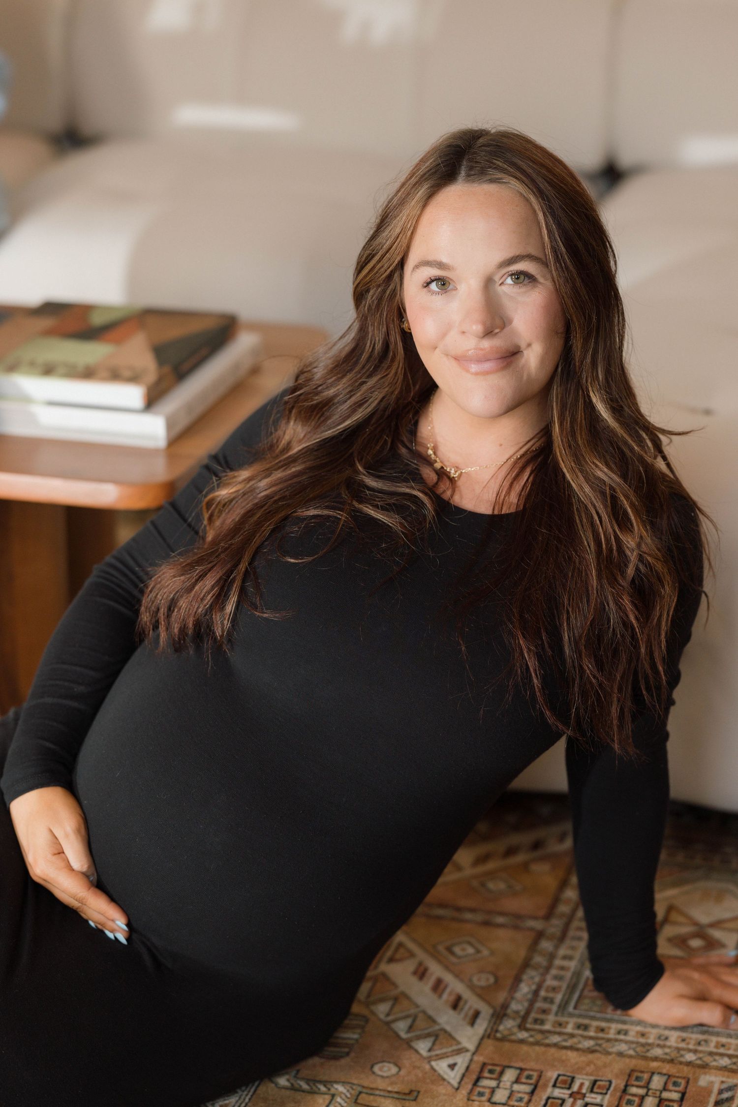 Pregnant woman with long brown hair in black dress sitting on patterned rug.