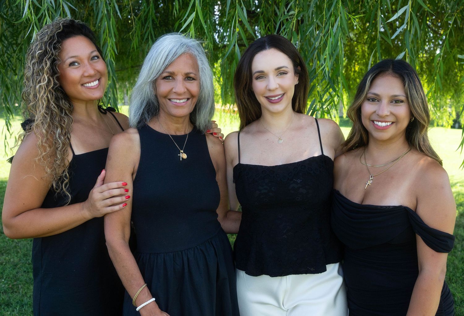 Four women in black outfits smiling together outdoors under a weeping willow tree on a sunny day.