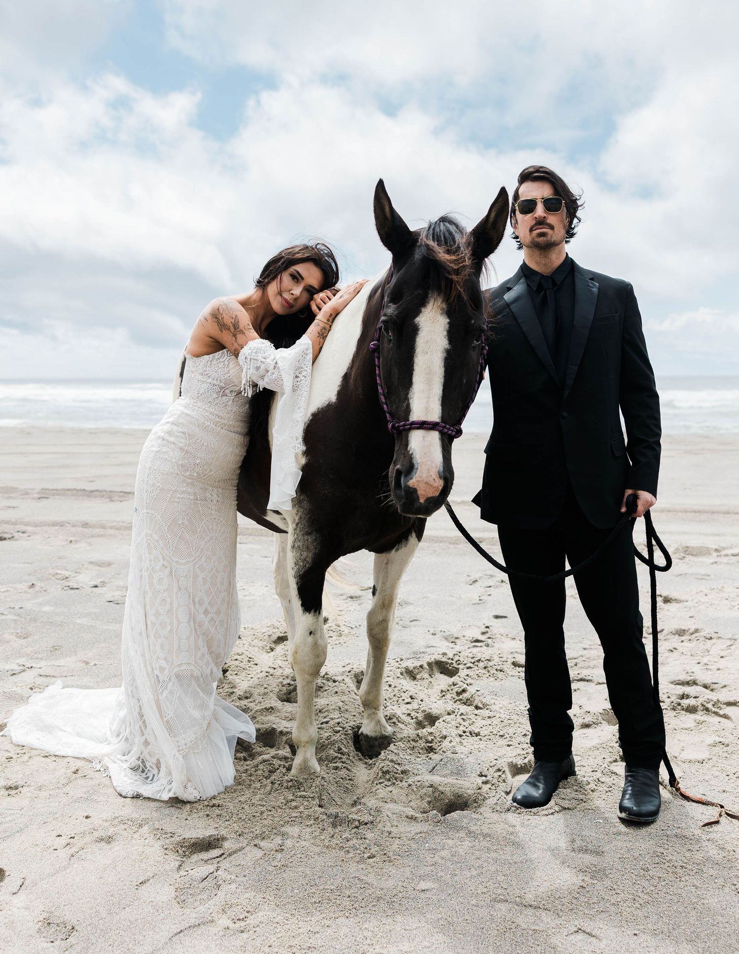 Bride and groom posing with a paint horse on a scenic sandy beach.