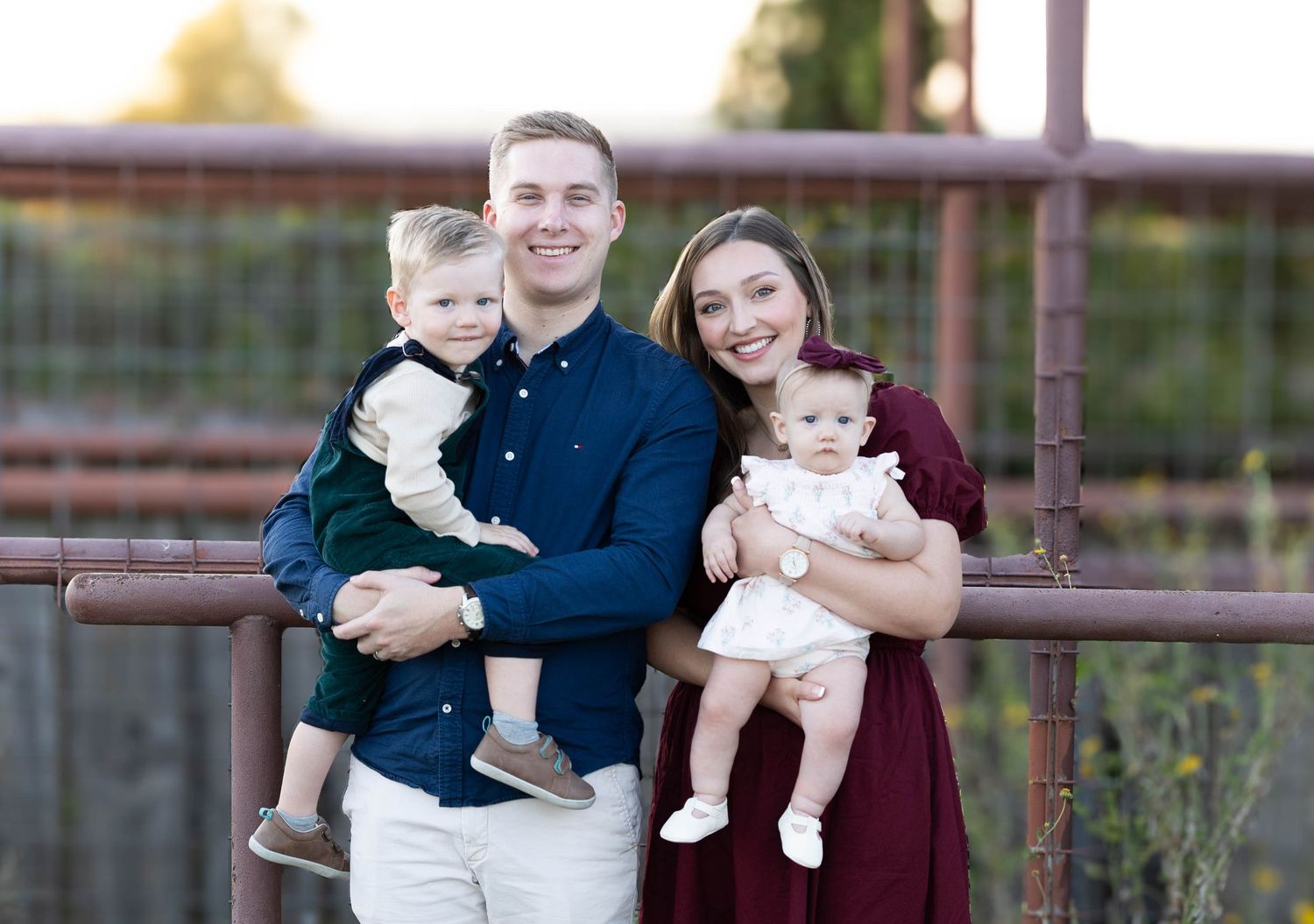 Young family of four posing outdoors, parents holding two small children, smiling at camera near rustic metal fence.