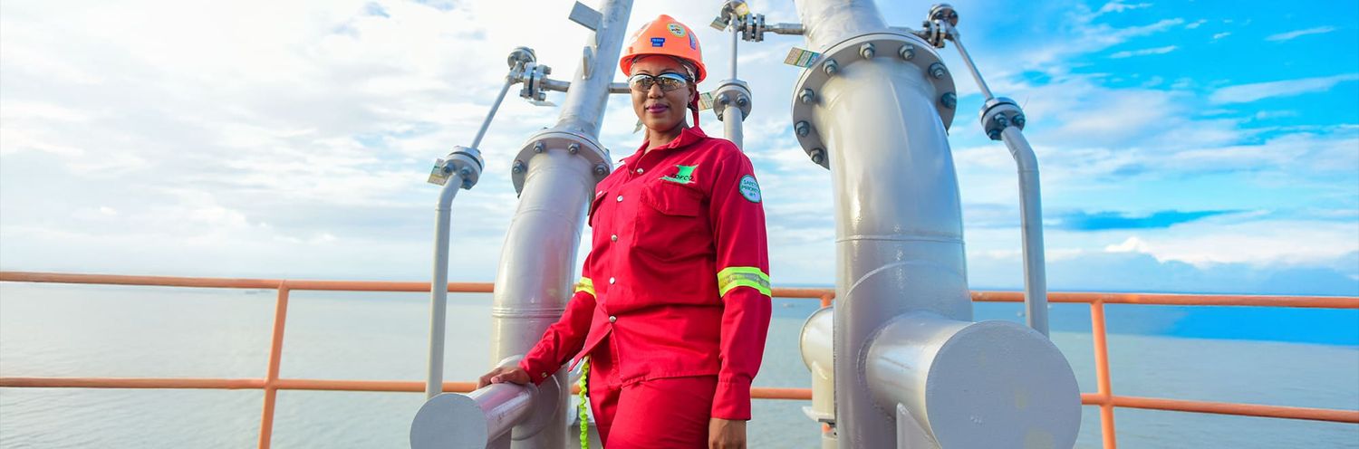 Worker in bright red coveralls stands between large industrial tanks on an offshore platform against blue sky.